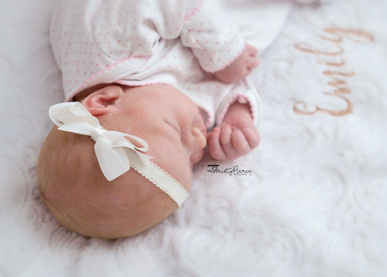 Sleeping newborn with white bow headband rests peacefully on soft white blanket embroidered with text.