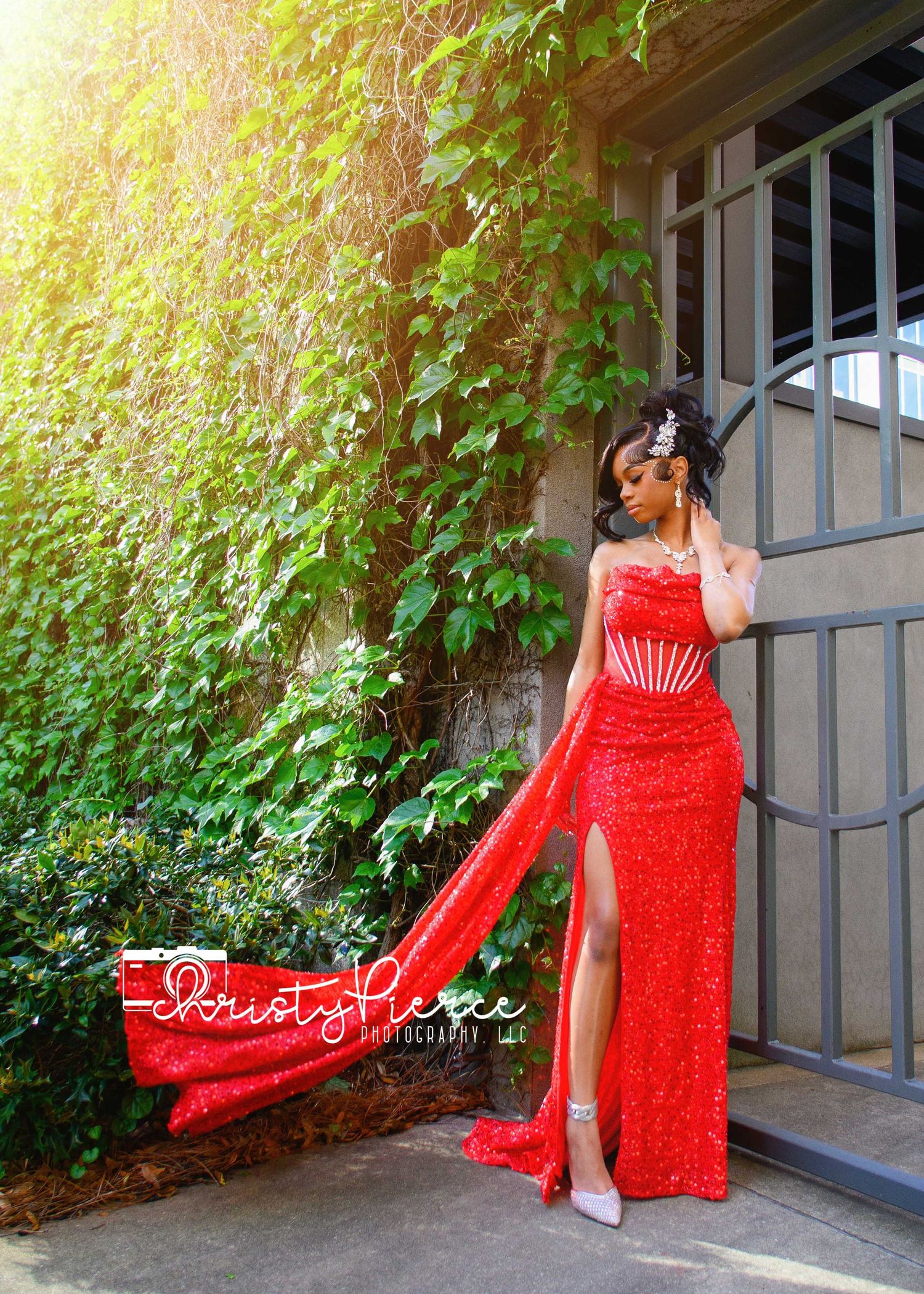 Woman in red sequin gown with slit poses outdoors near ivy wall, photographed by Dusty Pierce Photography.