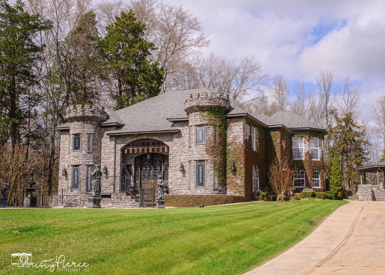 Luxury stone castle-style mansion with ivy-covered towers, arched entry, and lush green lawn surrounded by tall trees.
