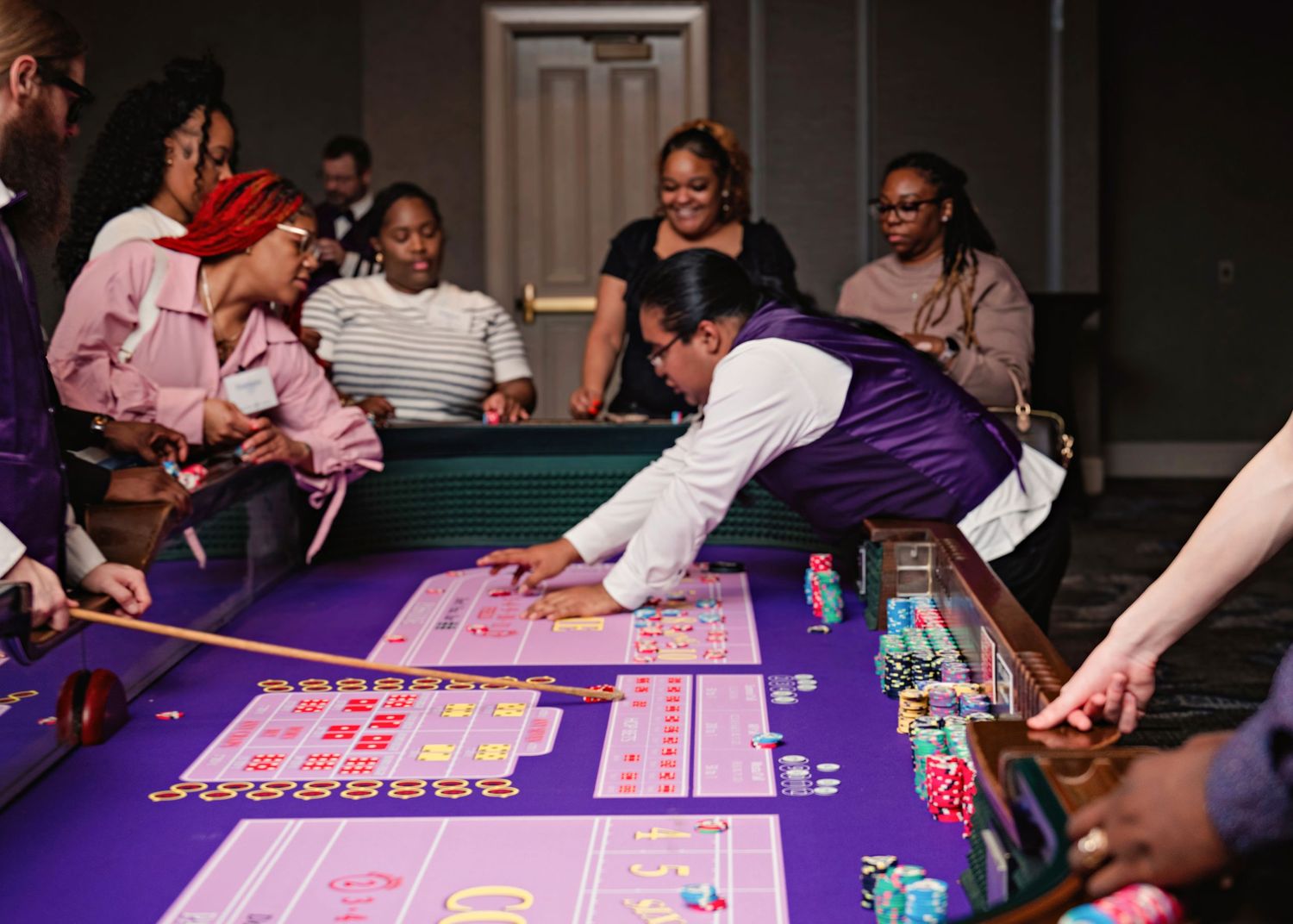 Players gather around a purple casino table placing bets and playing cards in a dimly lit gaming room.