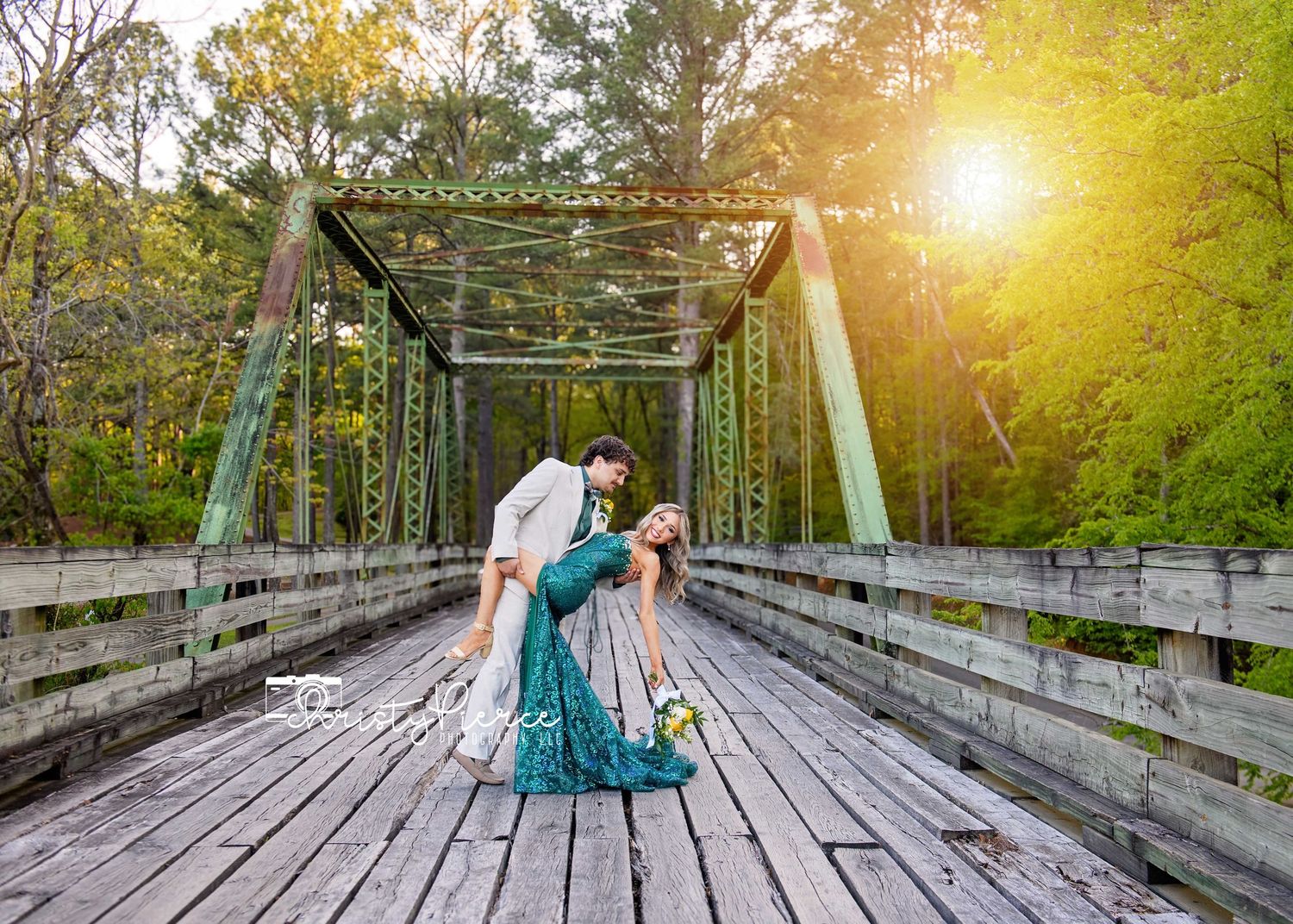 A couple in formal attire poses on a wooden bridge during golden hour sunset in a forest setting.