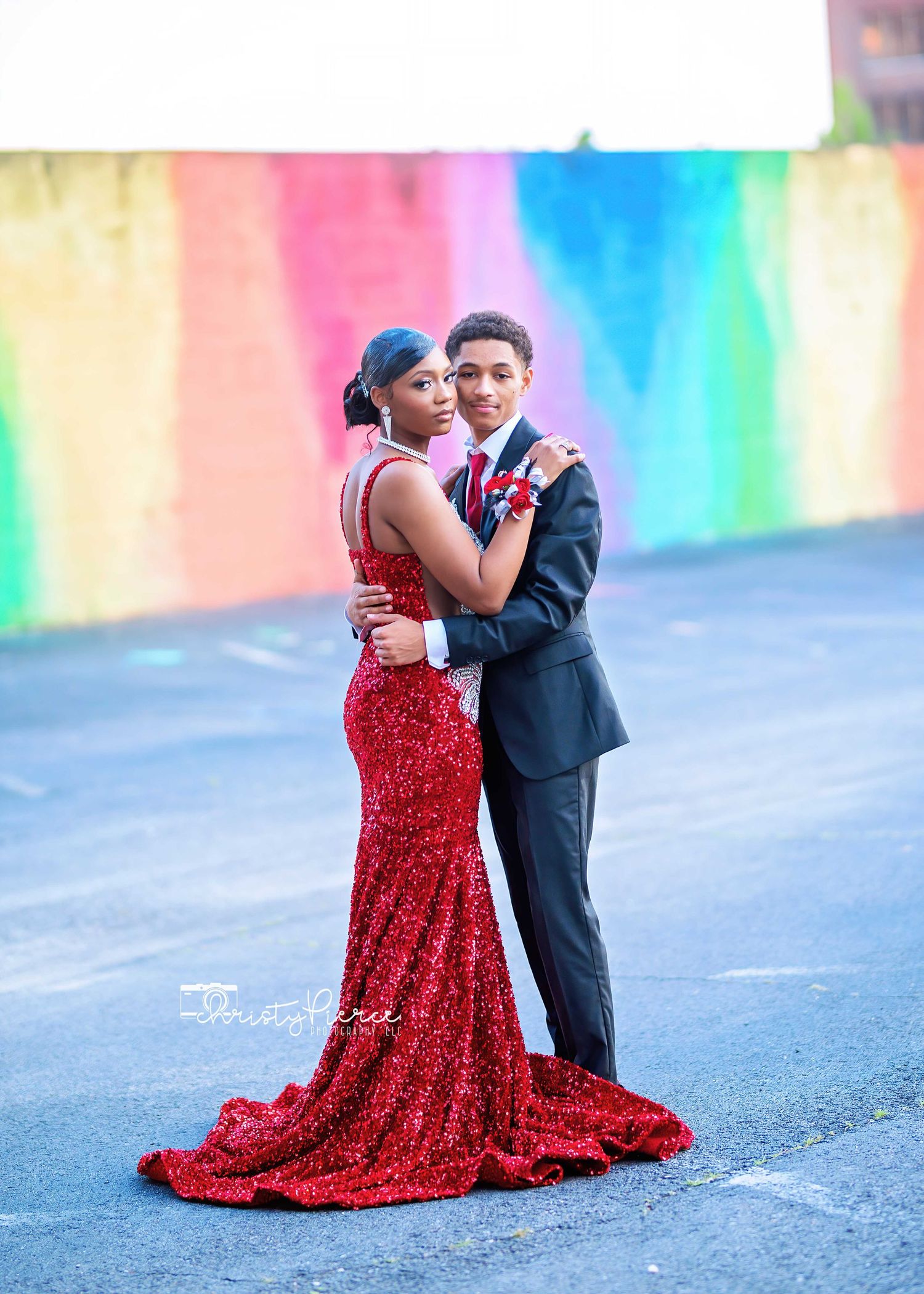 Couple posing against vibrant rainbow wall backdrop with person in red sequin gown and person in black suit.