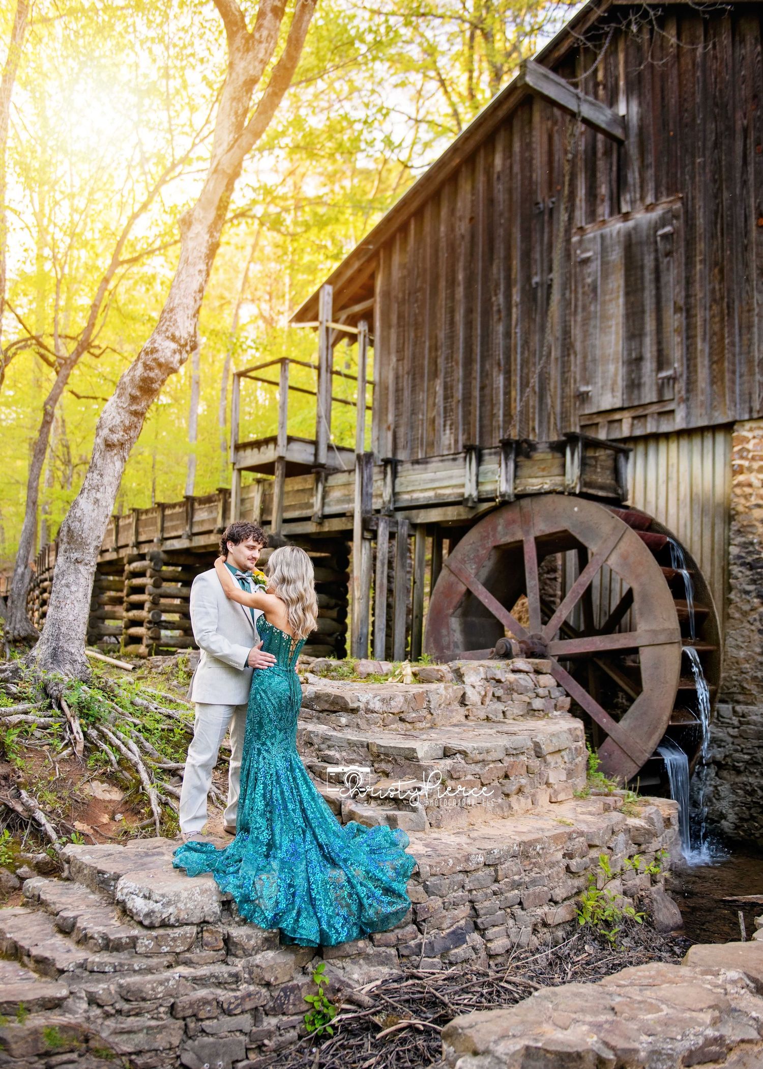 A rustic gristmill with wooden siding and large water wheel creates a romantic backdrop in golden afternoon sunlight.