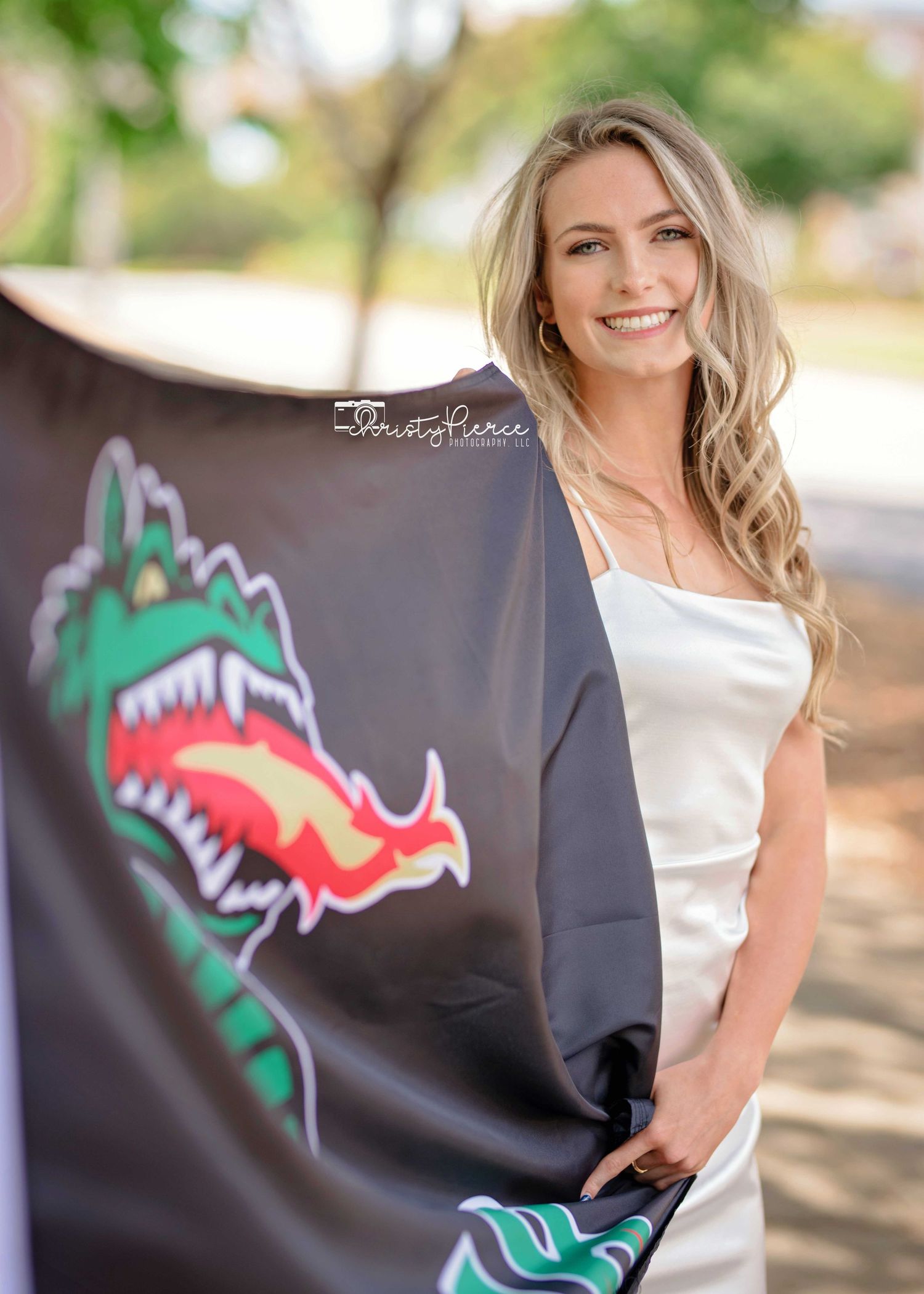 Smiling blonde woman in white dress holding a black dragon mascot flag outdoors at a graduation celebration.