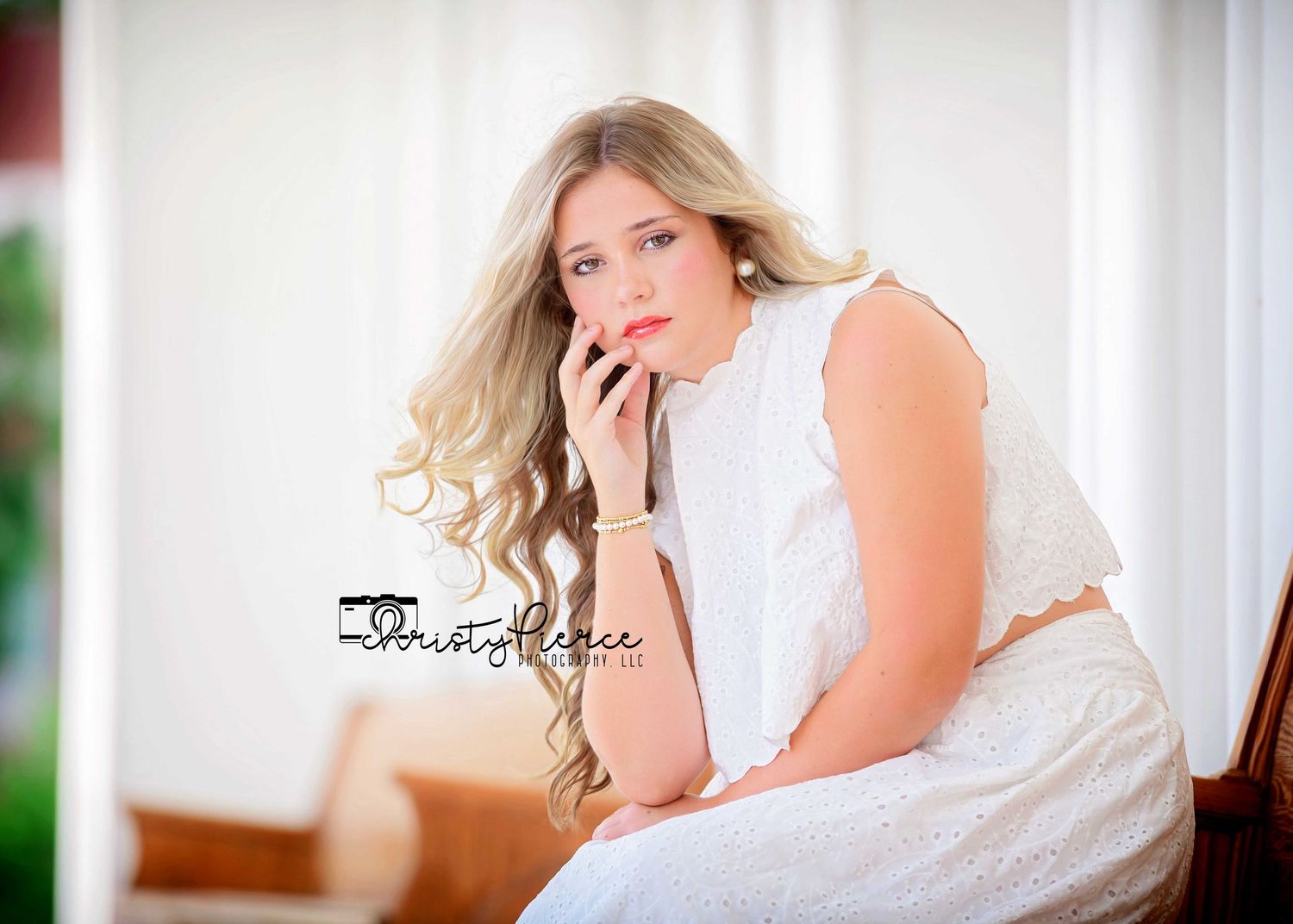 Young blonde woman in white sleeveless dress poses elegantly, resting chin on hand in a bright outdoor setting.