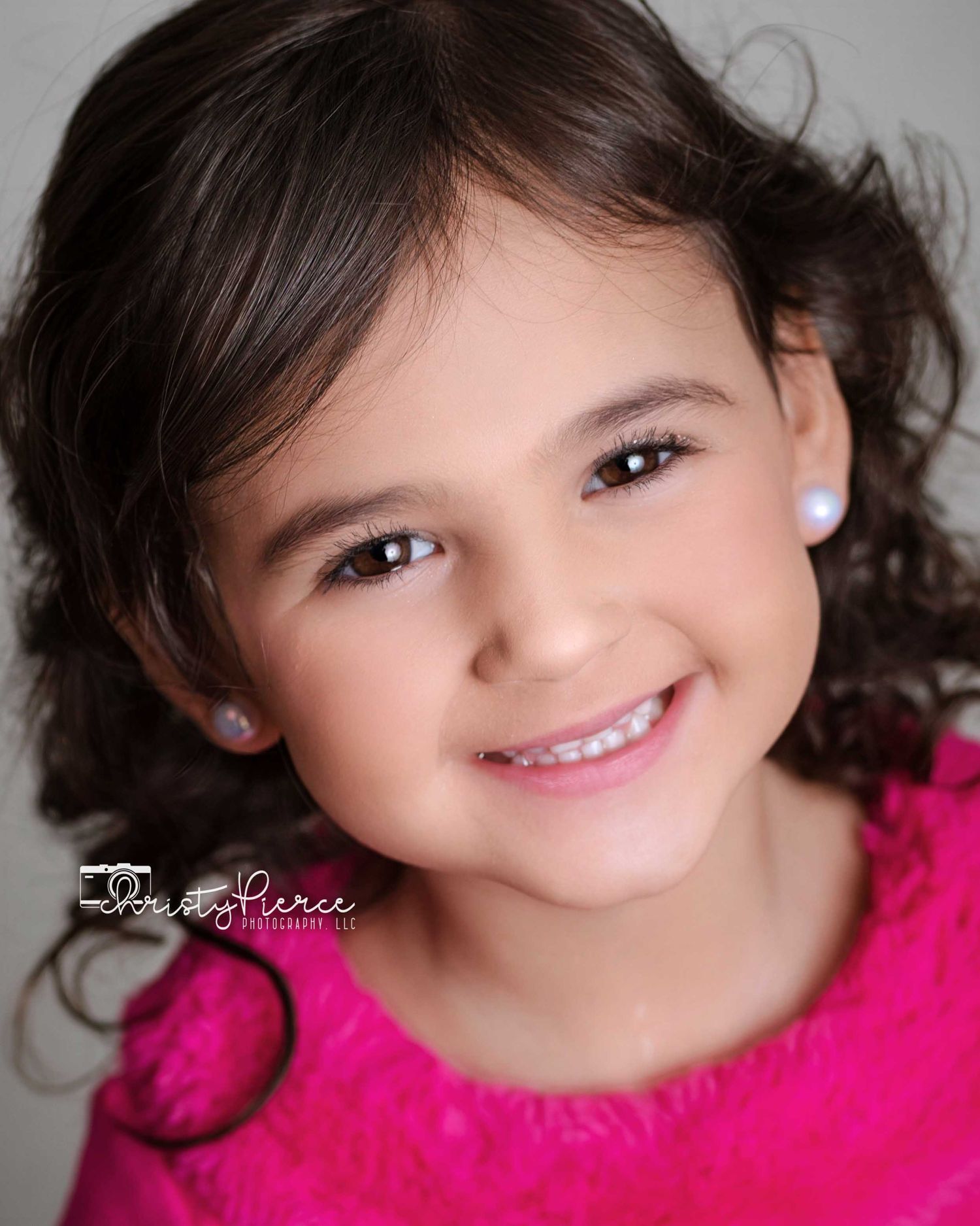 Child wearing bright pink top smiles warmly in professional portrait with soft lighting and dark curly hair.