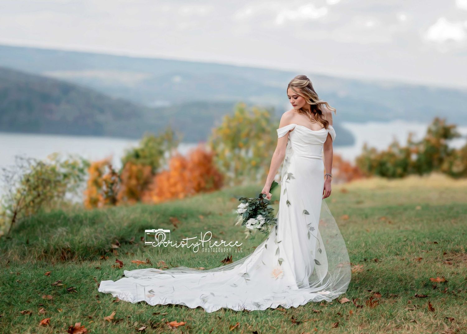 A bride in a flowing white lace gown stands on a hilltop overlooking an autumn lake on a misty afternoon.