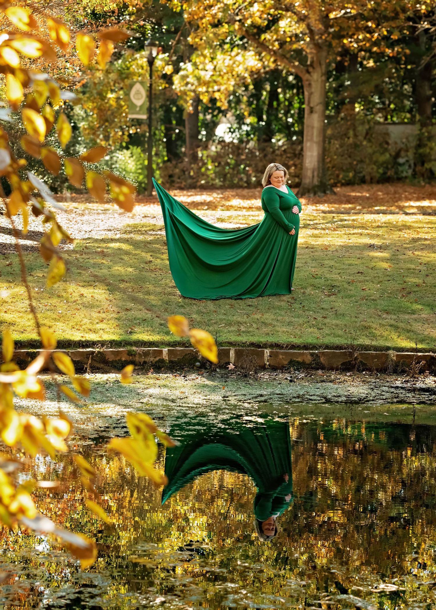 A figure in an emerald green flowing dress creates artistic reflections while posing near a tranquil autumn pond.