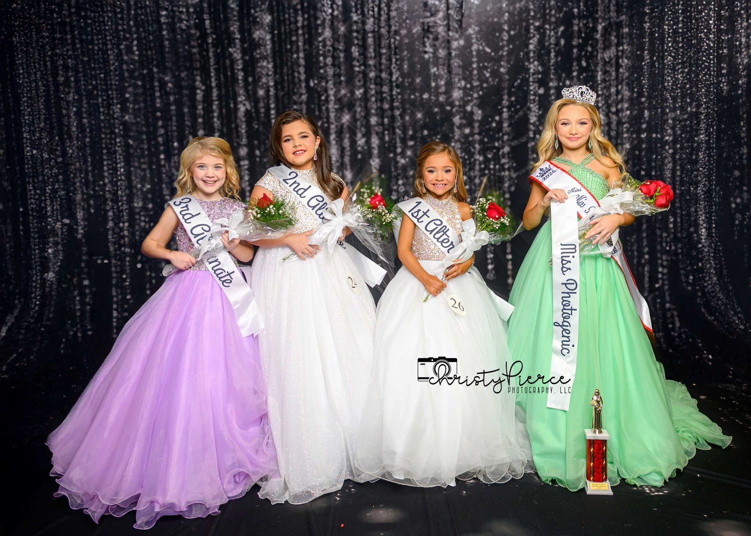 Four pageant contestants wearing elegant ball gowns and crowns pose together against a sparkly black backdrop.