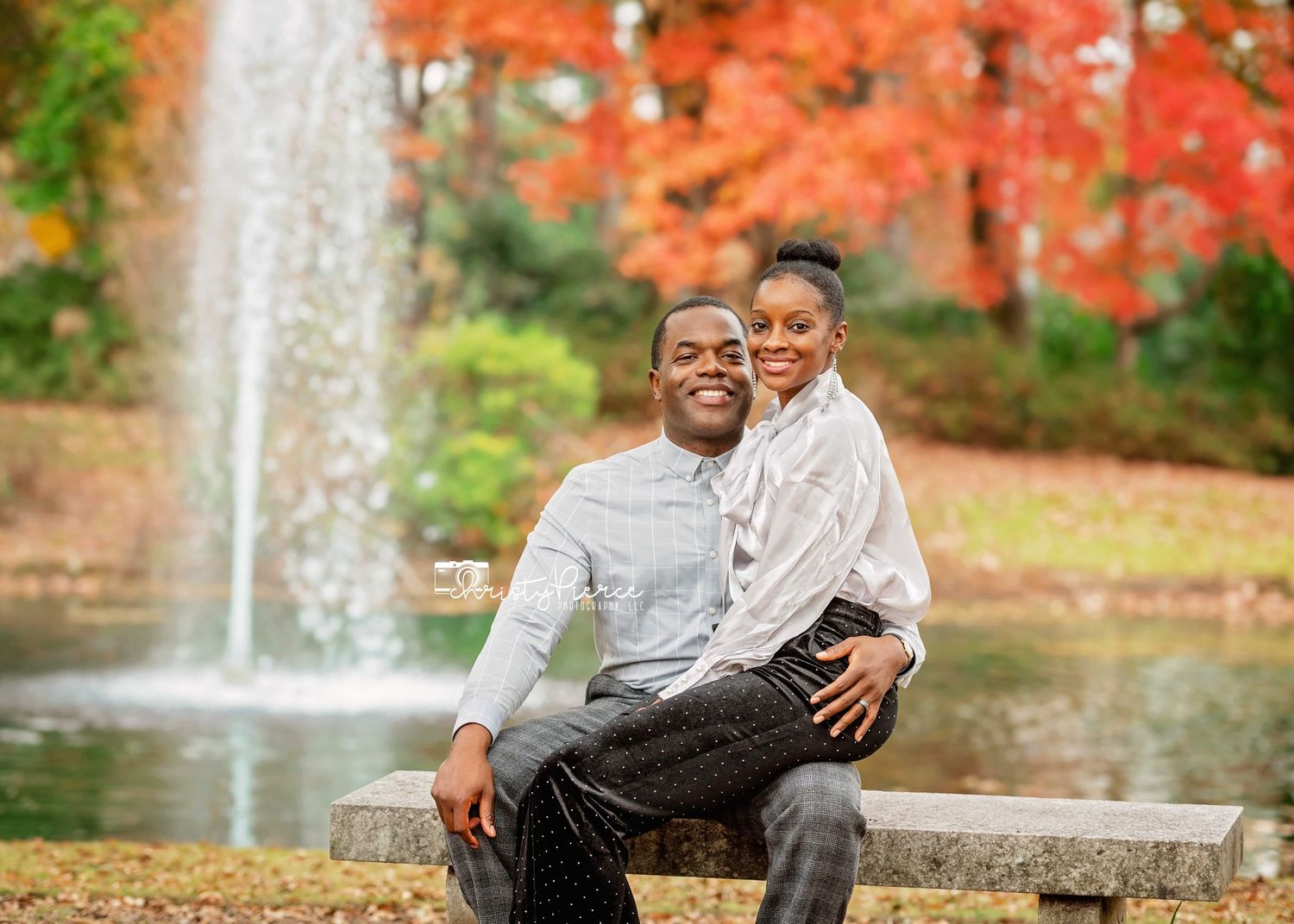 A couple sits together on a bench surrounded by vibrant autumn foliage and a peaceful water fountain in the background.