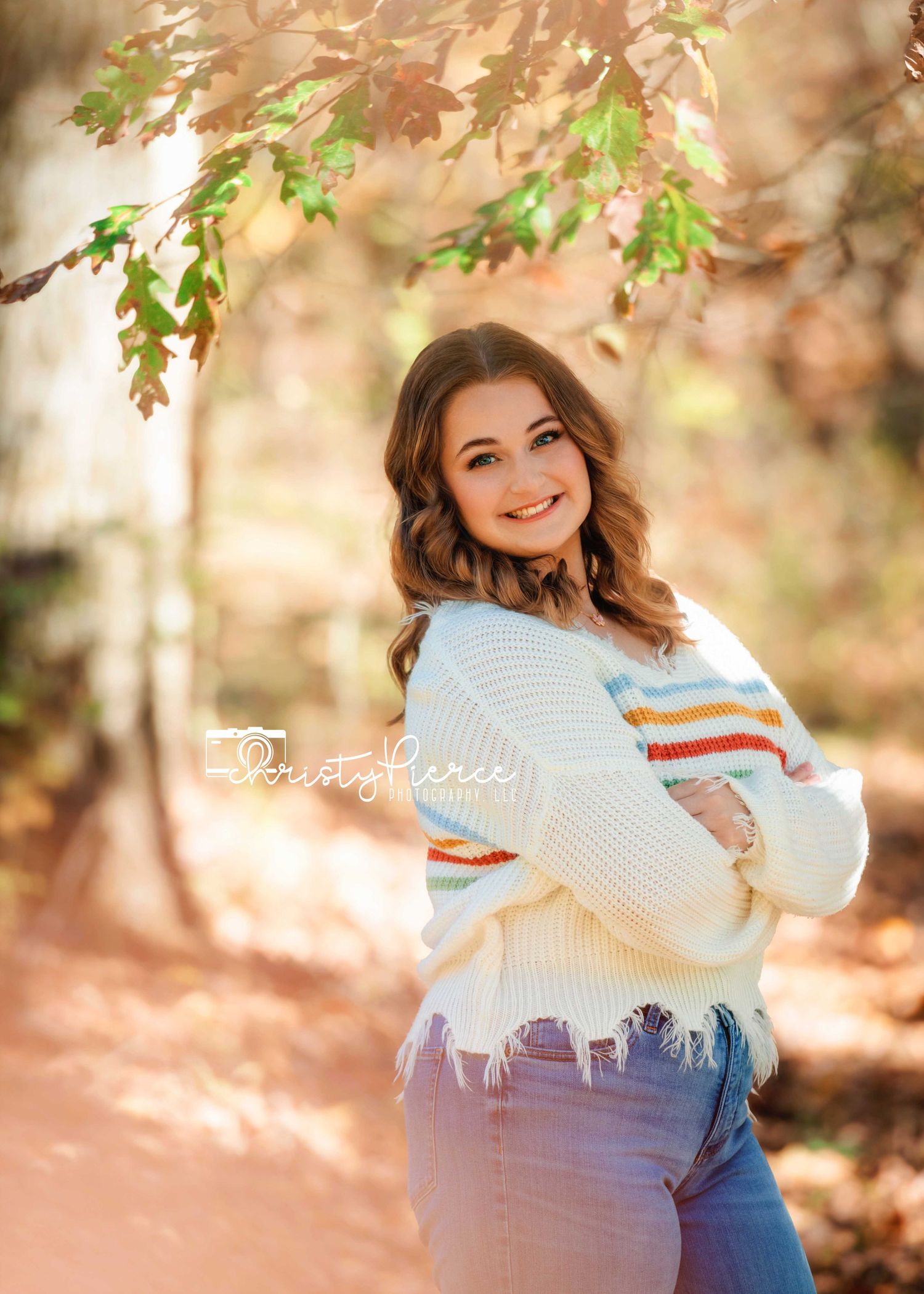 A smiling young woman in a cream striped sweater and jeans poses outdoors among autumn foliage in a warm forest setting.