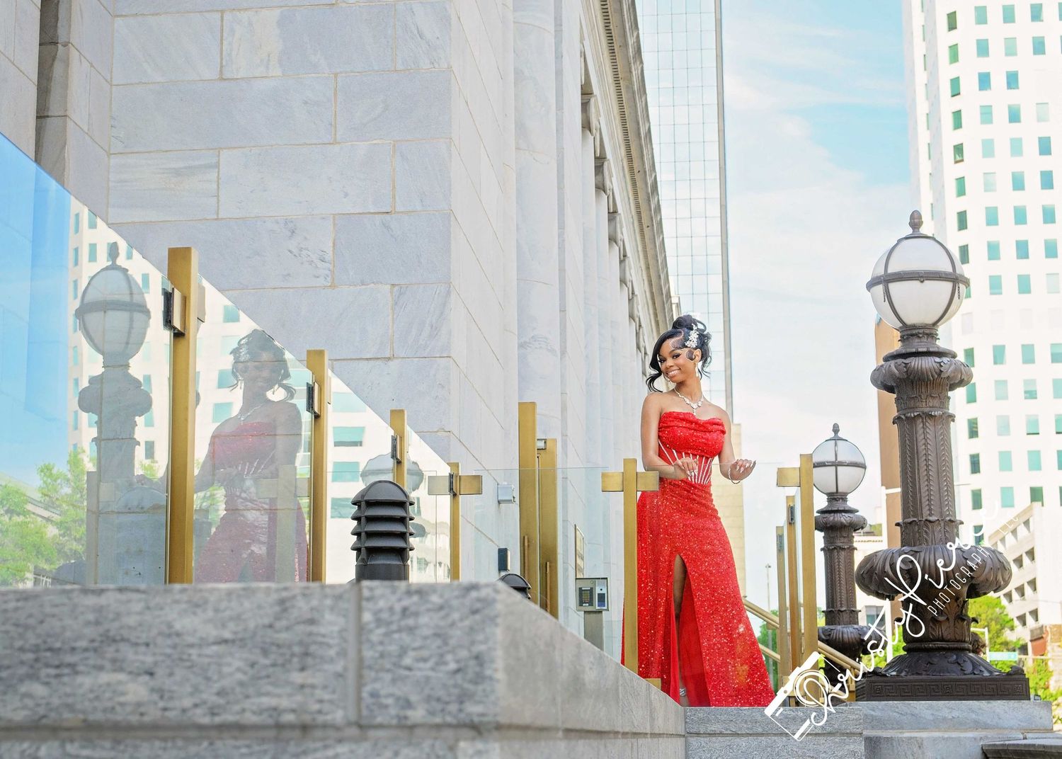 Woman in red off-shoulder gown poses on grand steps with gold lamp posts and modern buildings in background.