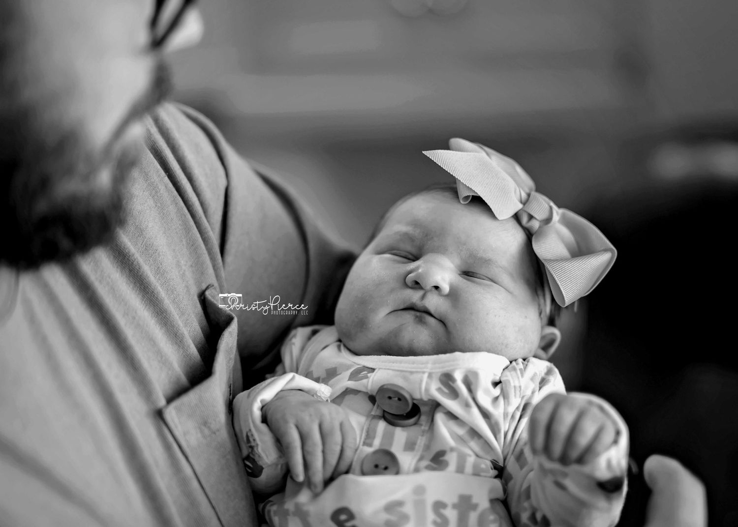 A peaceful sleeping newborn wearing a bow headband rests against someone's shoulder in black and white.
