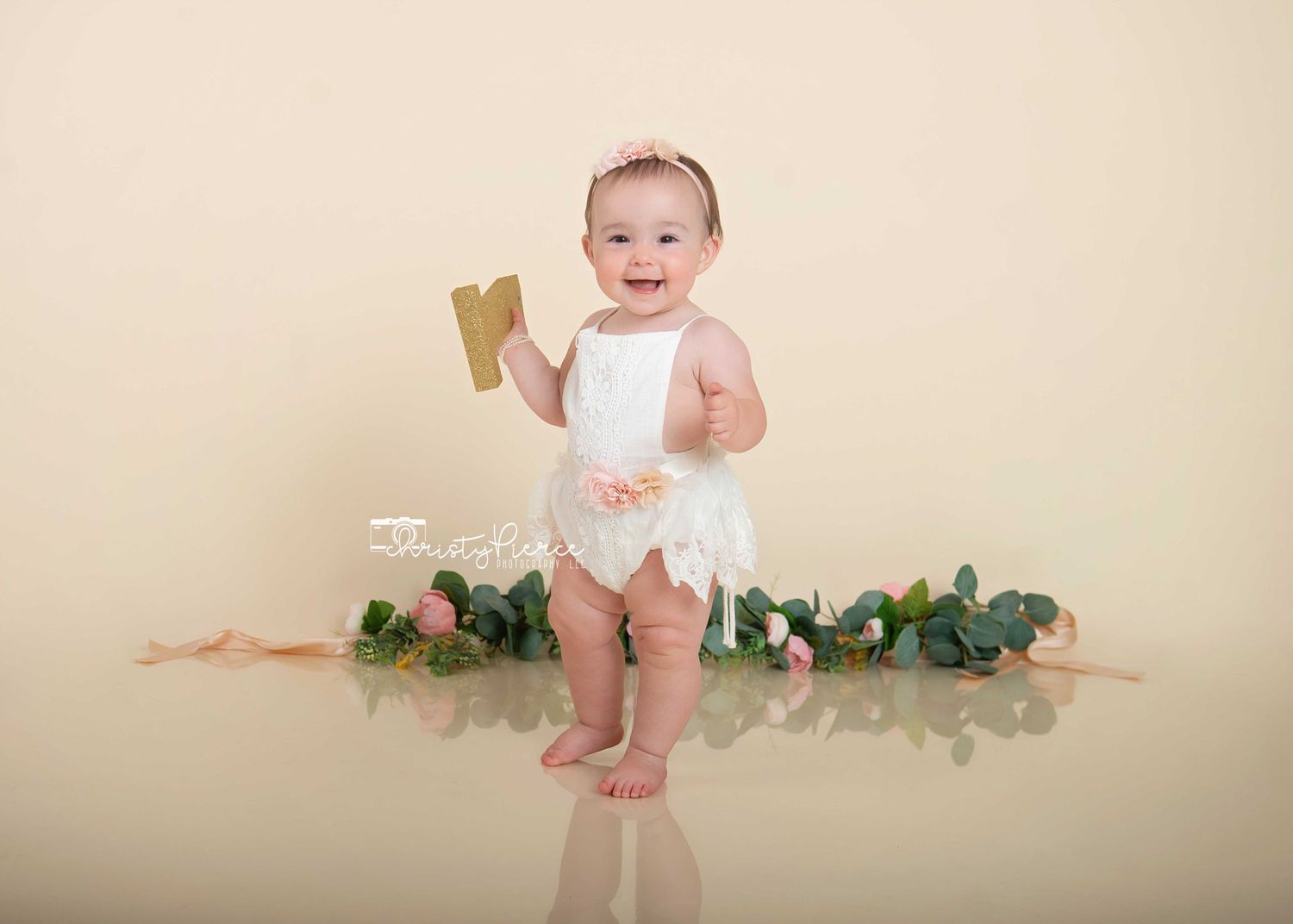 A happy baby in a white tutu stands in front of greenery and floral decorations on a beige backdrop.
