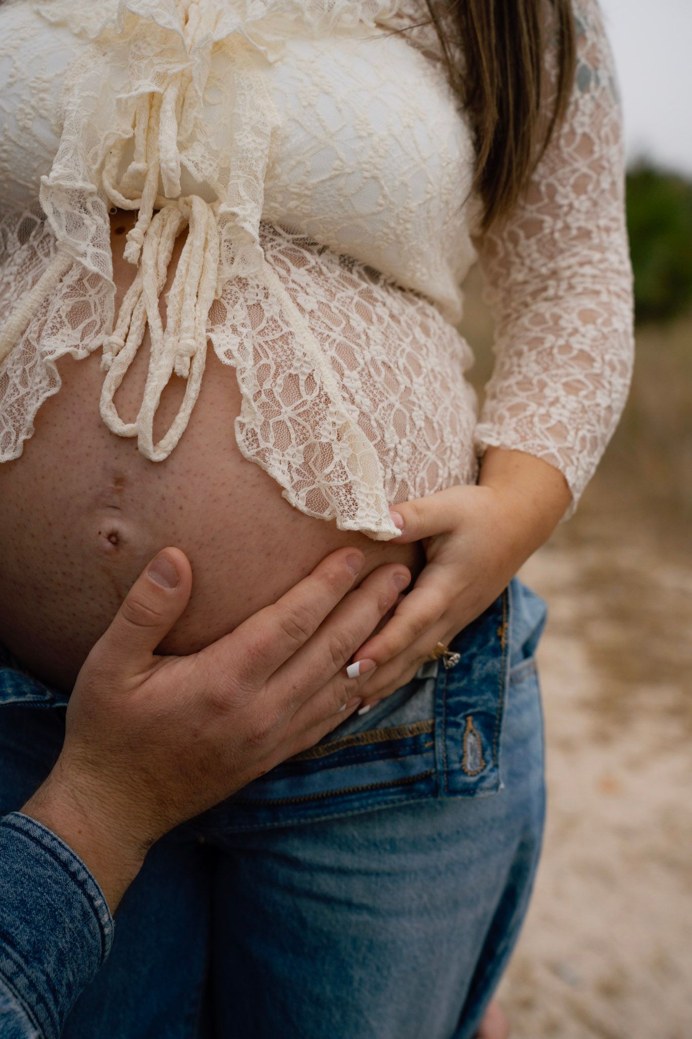 A pregnant belly wrapped in delicate white lace is tenderly touched by two hands against a blurred outdoor background.