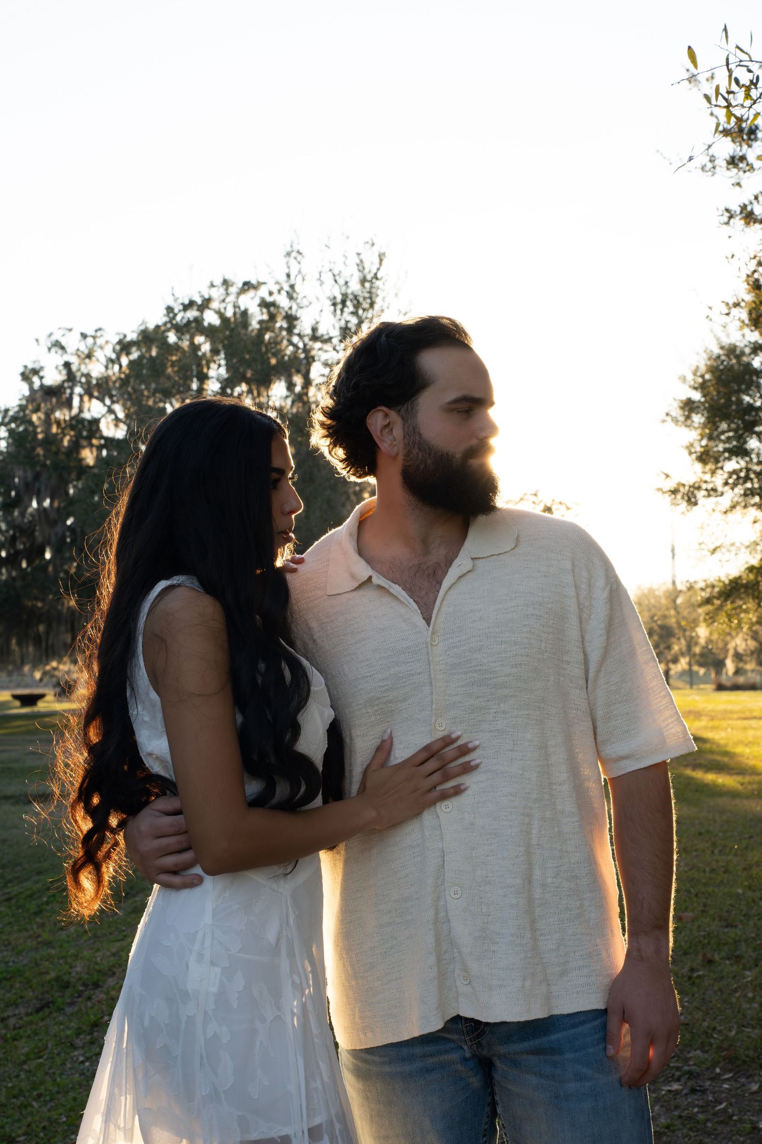 A couple embraces in a sunlit farm during golden hour wearing white and denim casual attire during Engagement Session at Congaree and Penn.