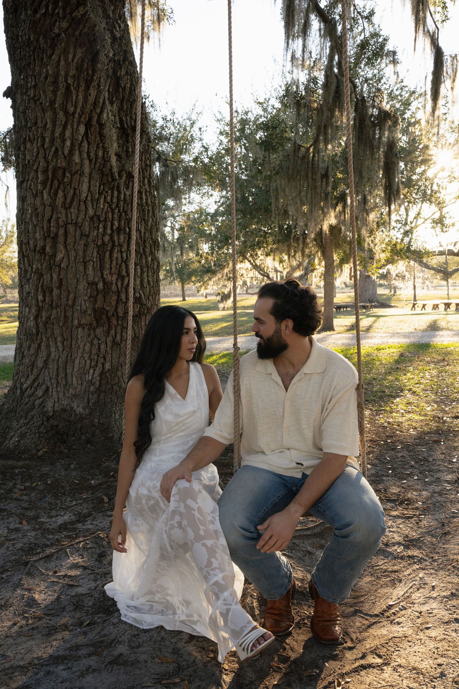 A couple sits together under Spanish moss-draped trees in a romantic outdoor setting at sunset.