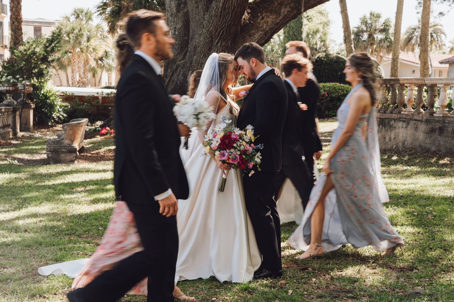 Wedding party members gather under trees in a park during an outdoor celebration.