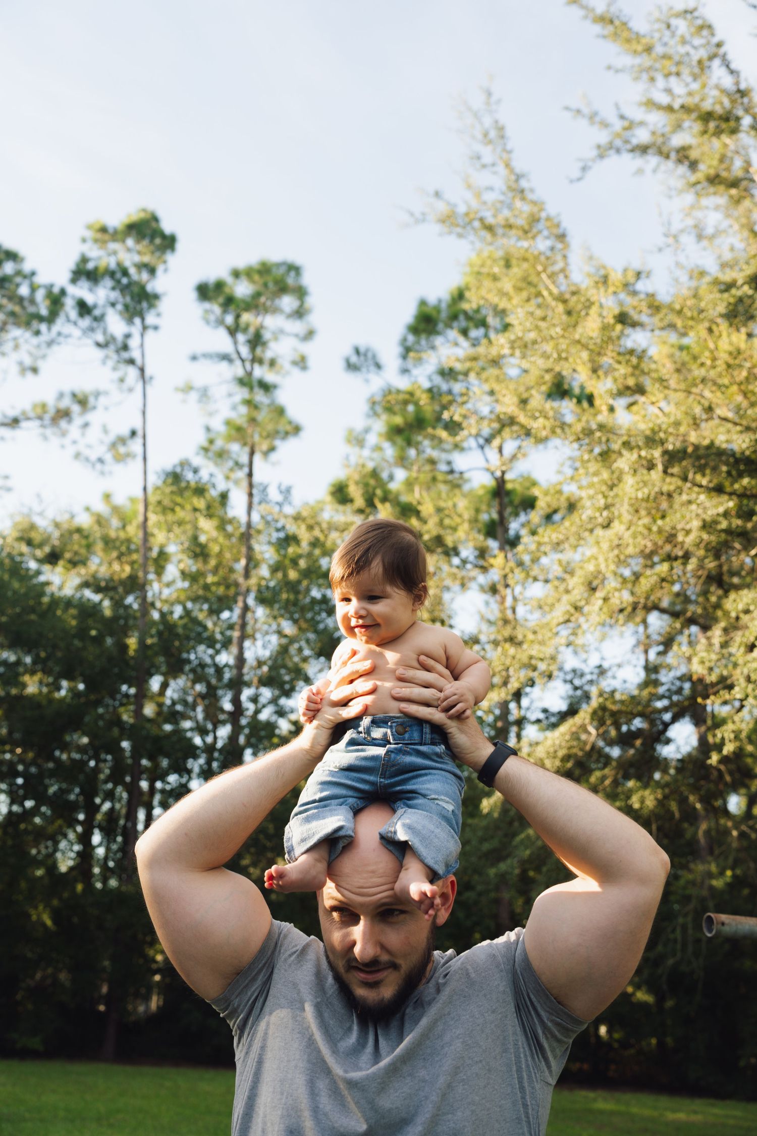 A playful outdoor scene showing someone lifting a small child against a backdrop of trees and sunlit foliage.
