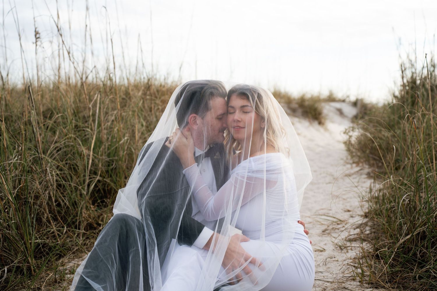 A romantic couple embraces under a white veil on a sandy beach path surrounded by coastal grass.