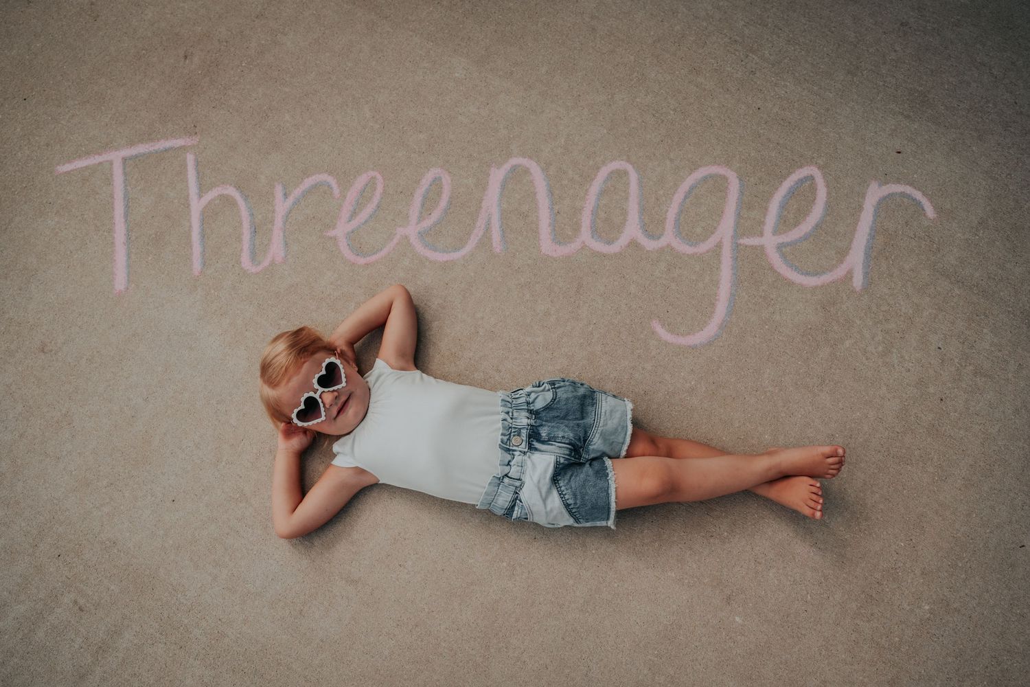 A person lies on the floor beneath the word 'Threenager' written in chalk on a beige carpet.