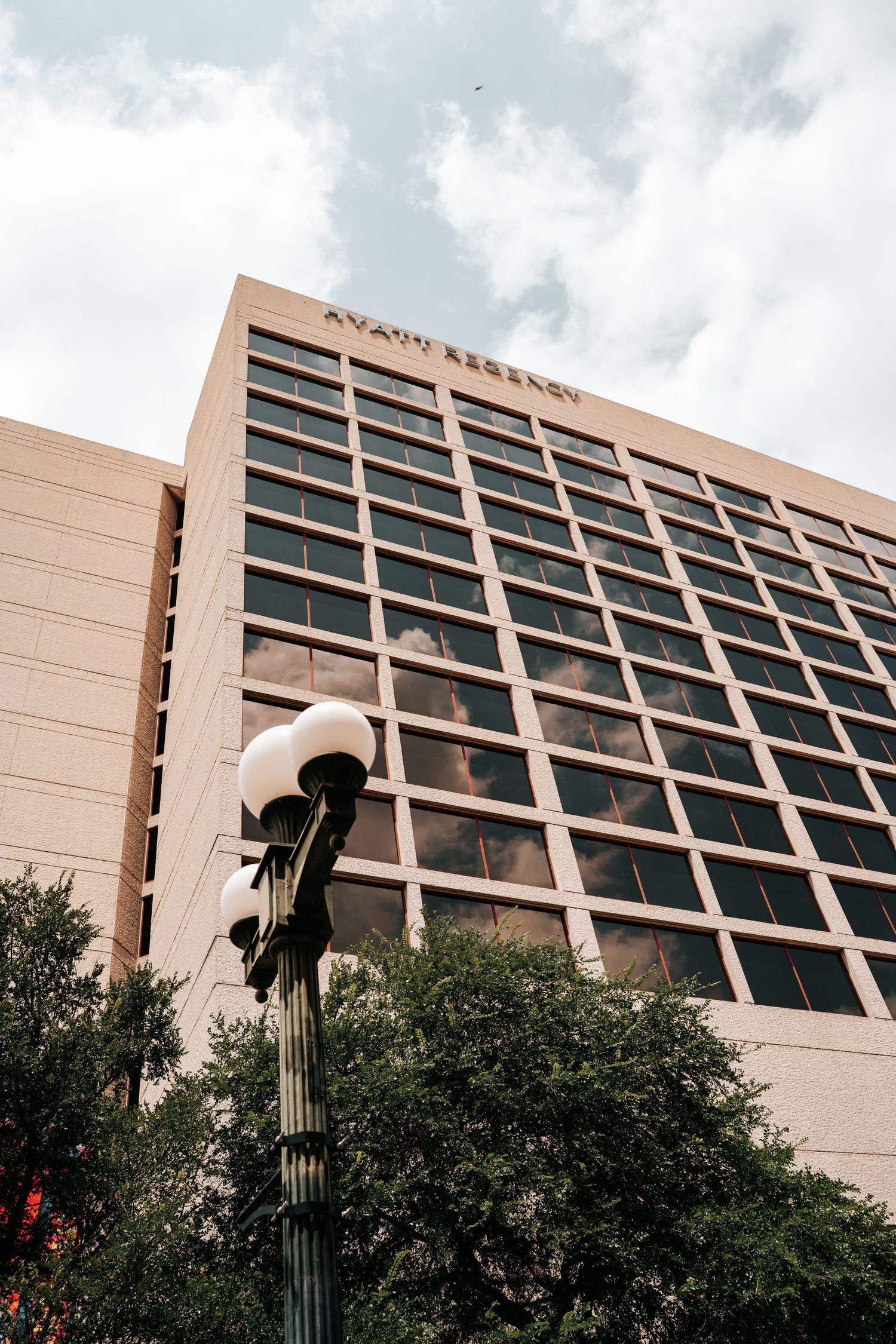 Hyatt Regency hotel building exterior with grid windows, street lamp, and trees against cloudy sky.