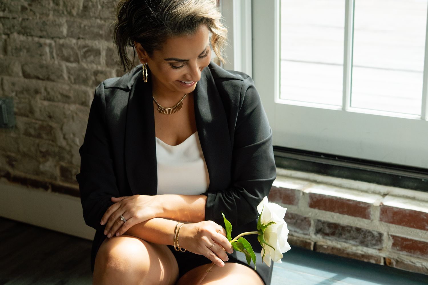 Woman in black blazer and white top sits contemplatively holding a white flower near a brick wall window.