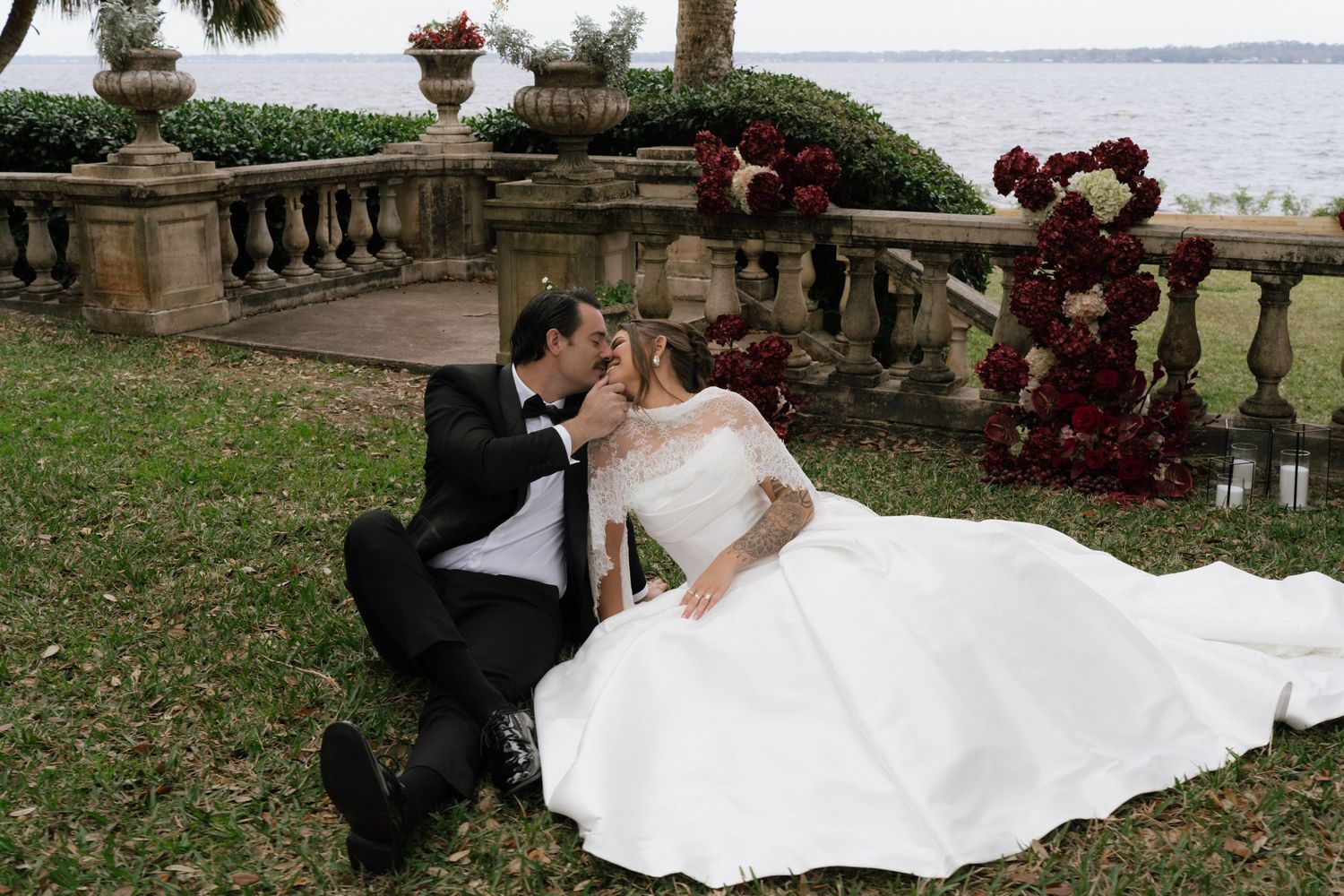 A couple in wedding attire shares a romantic moment on a stone balcony overlooking a waterfront garden.