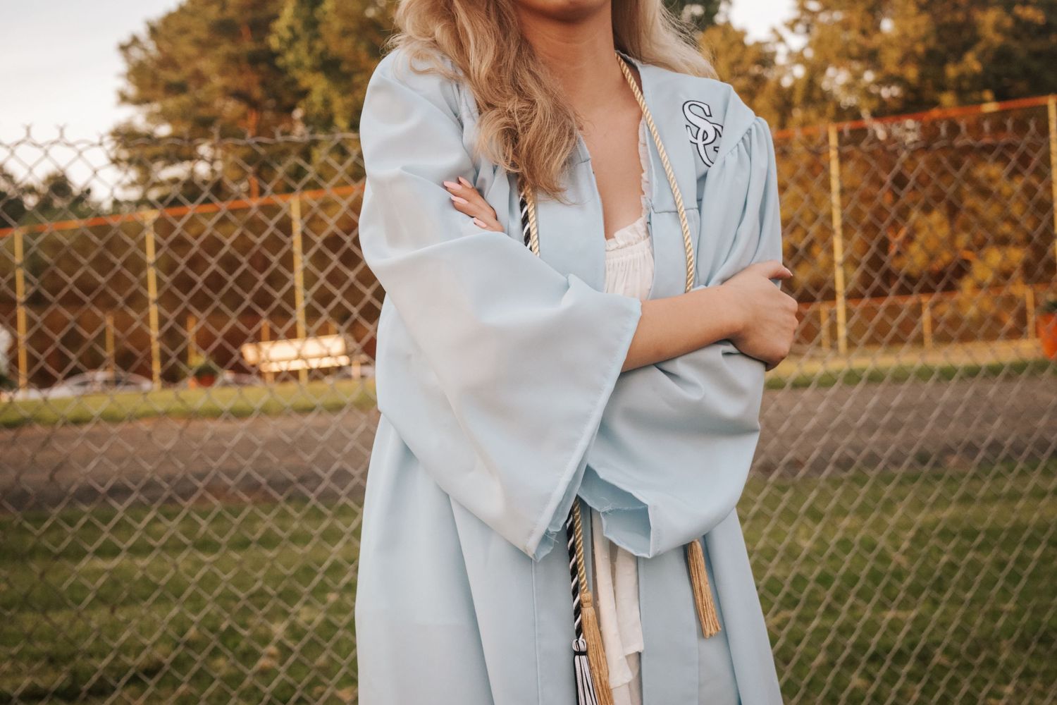 A person wearing a light blue graduation gown stands in front of a chain link fence at sunset.