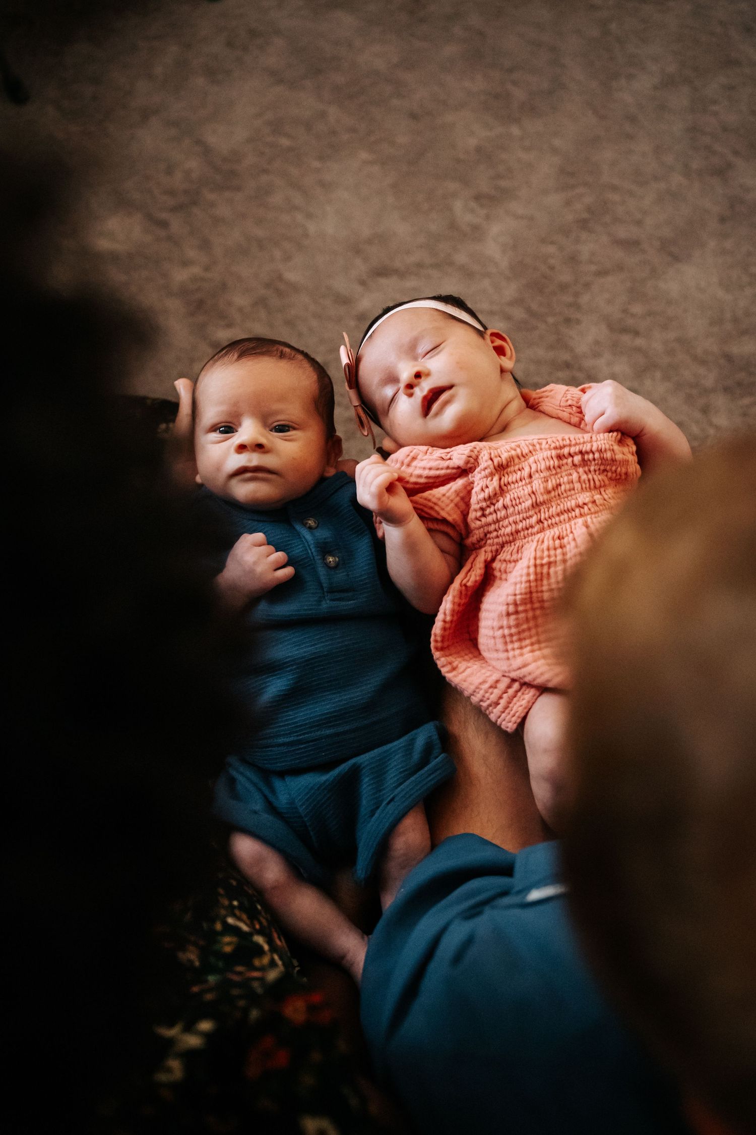 Two babies lying together, one in blue and one in coral colored outfits.