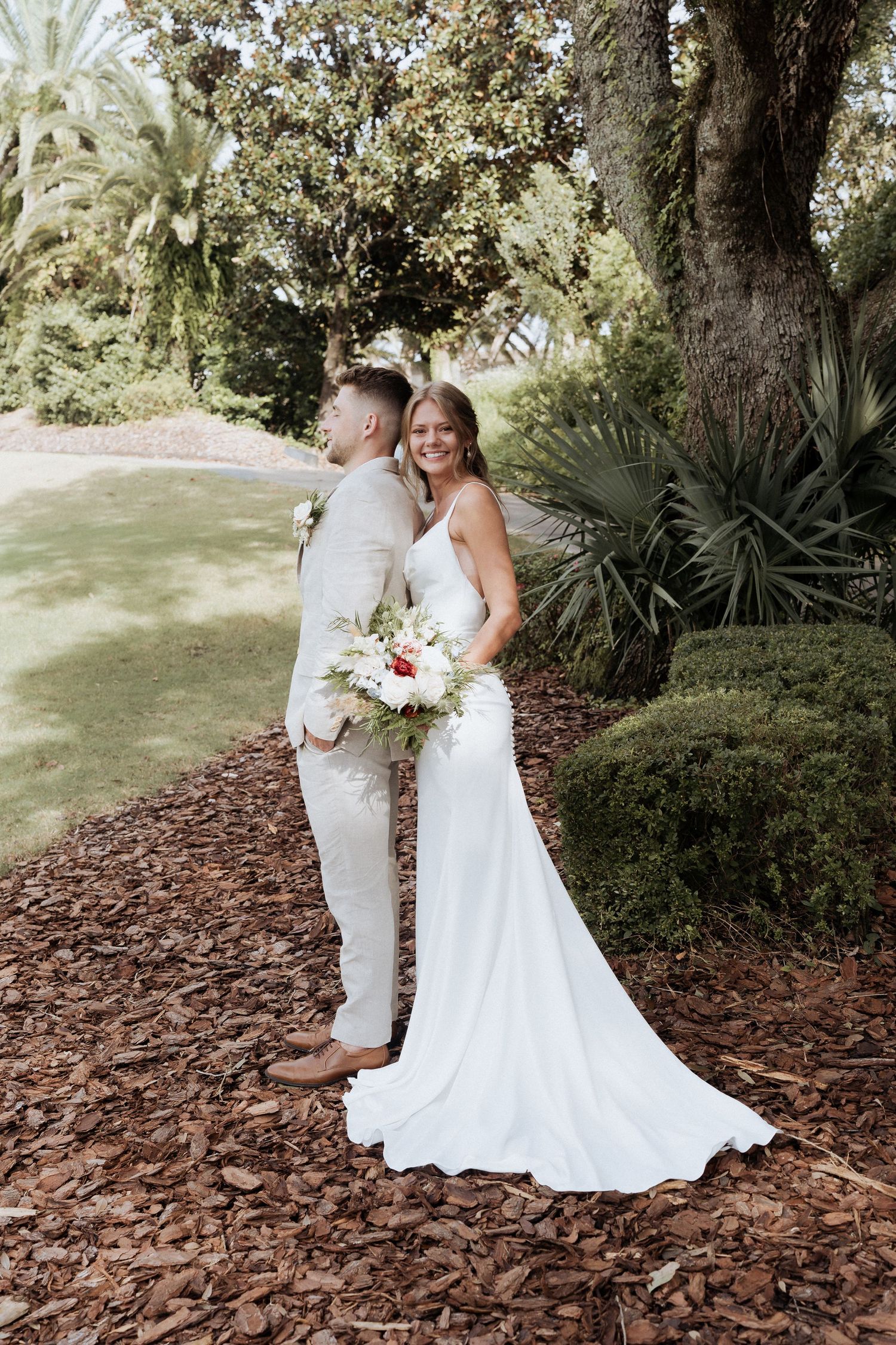A romantic outdoor wedding photoshoot in a garden setting with two people in white dresses among trees and mulch.