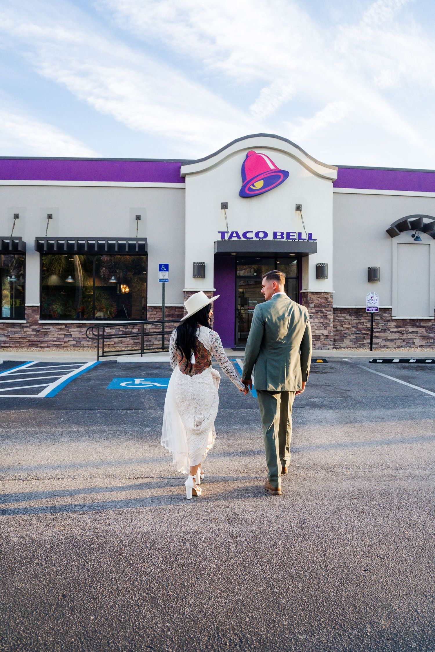 A couple in formal attire walks hand in hand towards a Taco Bell restaurant entrance under a blue sky.