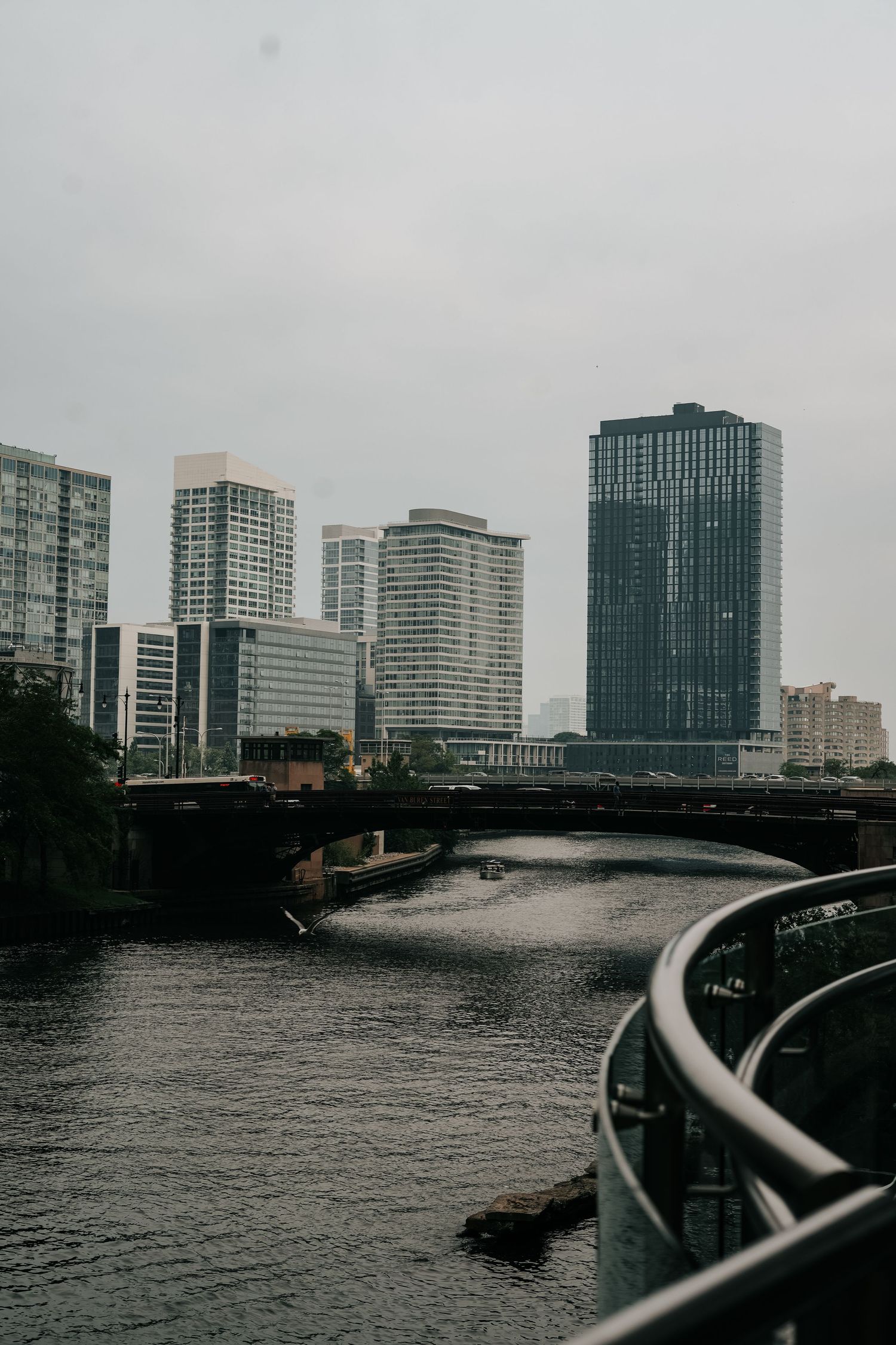 Modern city skyline with tall glass buildings reflected in river water along curved railing on cloudy day.