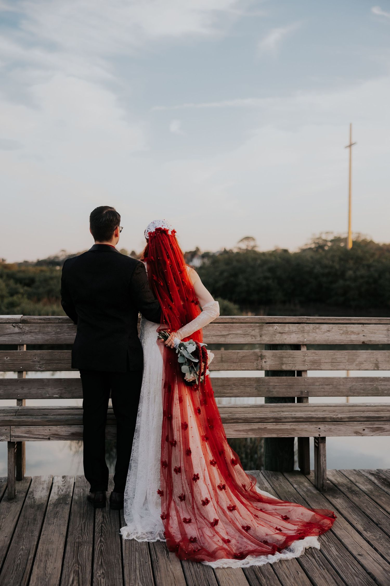 A couple in wedding attire sits together on a wooden dock overlooking a lake at sunset.