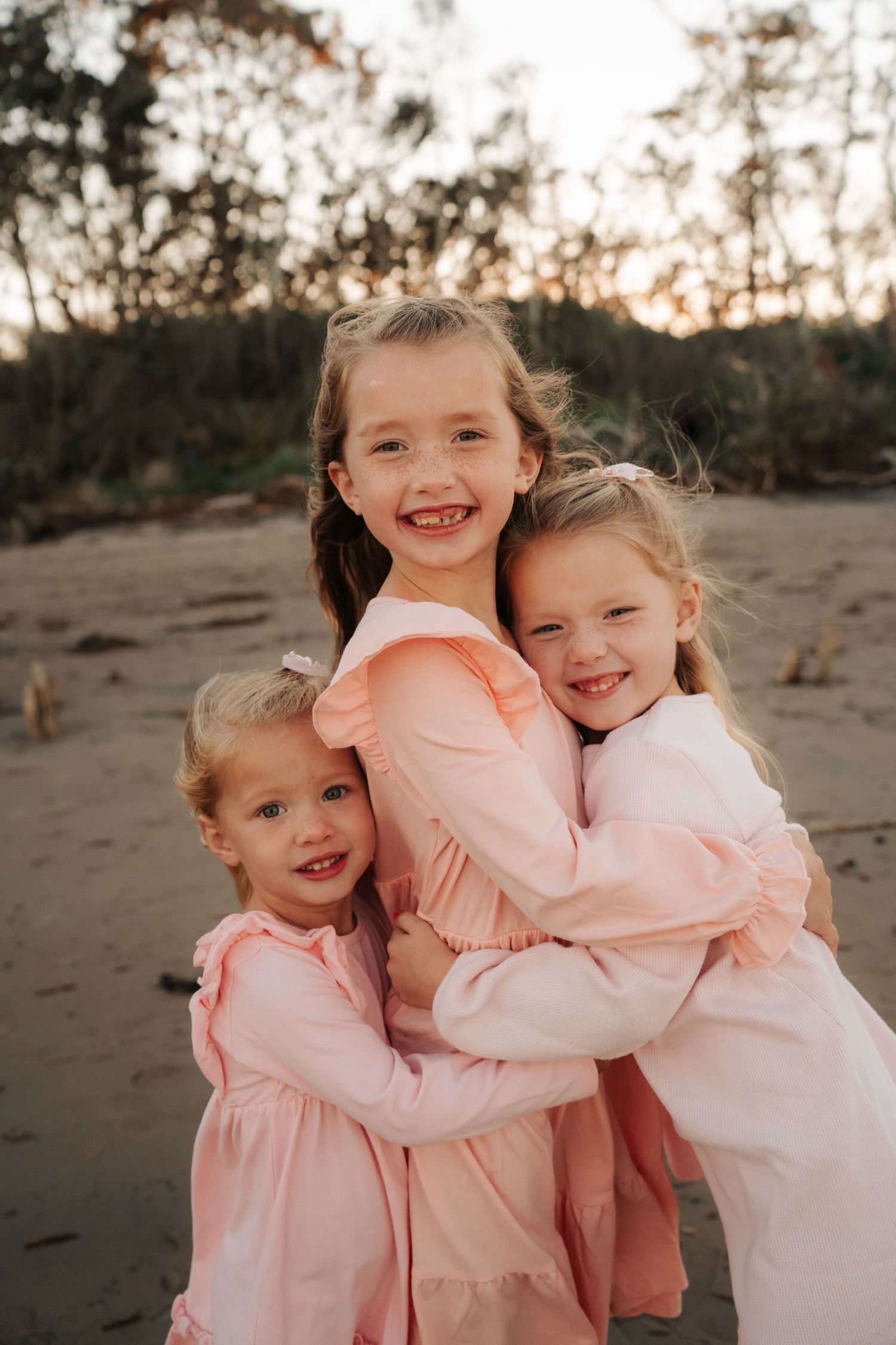 Three young girls in matching pink dresses hugging and smiling on beach.