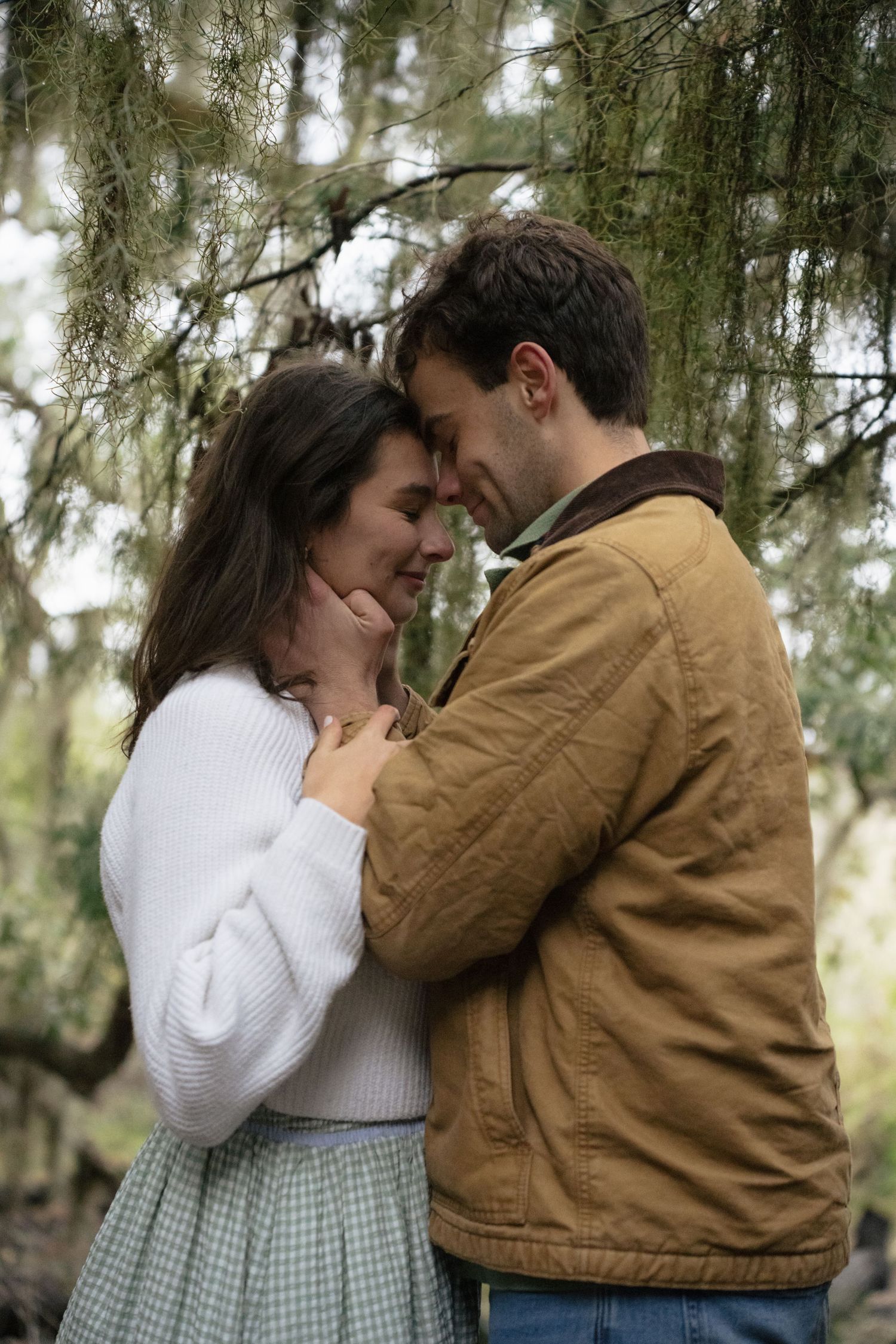 A romantic couple embraces tenderly beneath evergreen trees, with her wearing a white sweater and plaid skirt.