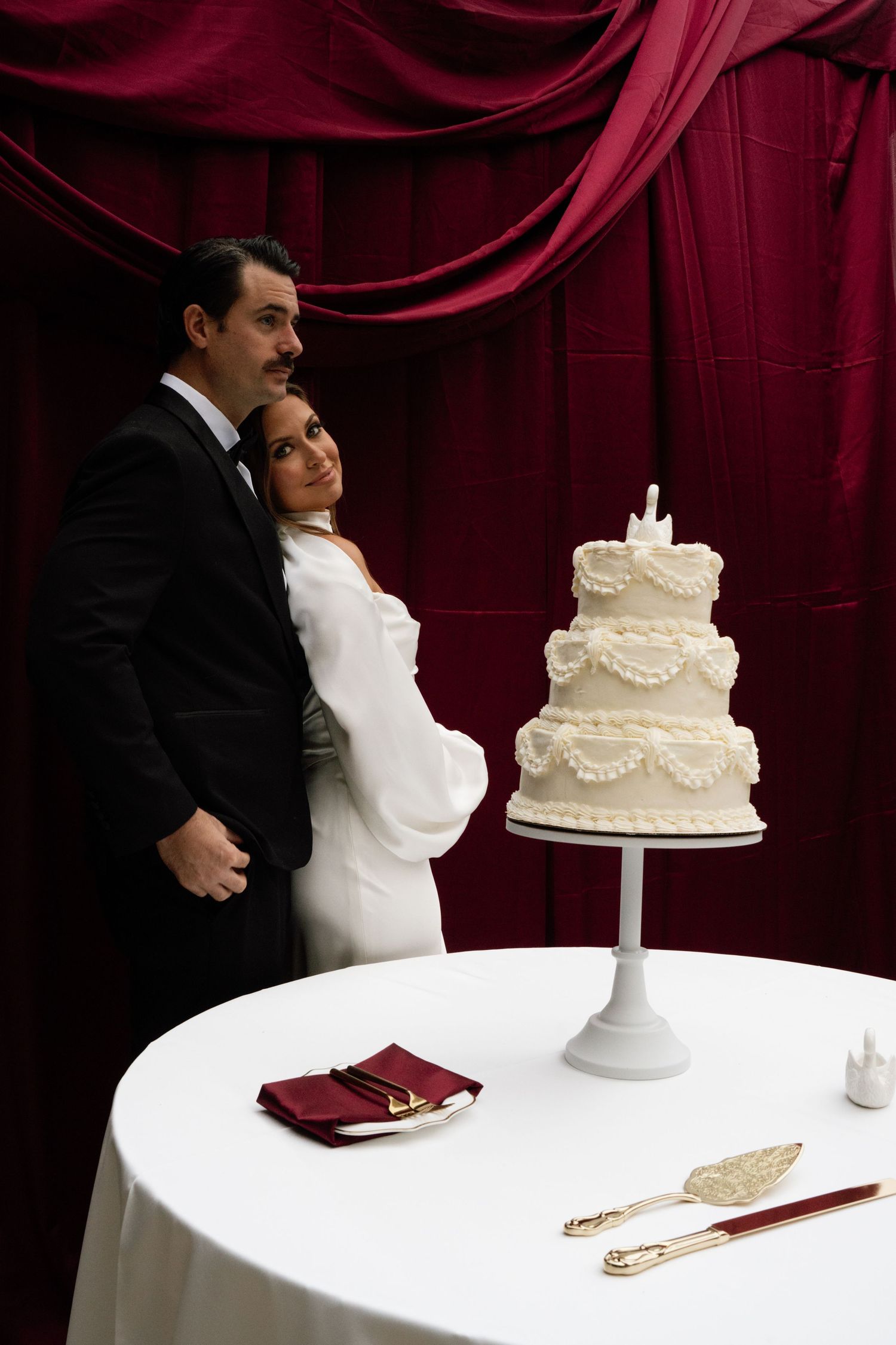 A wedding couple stands near an elegant white tiered cake against a deep red velvet backdrop.