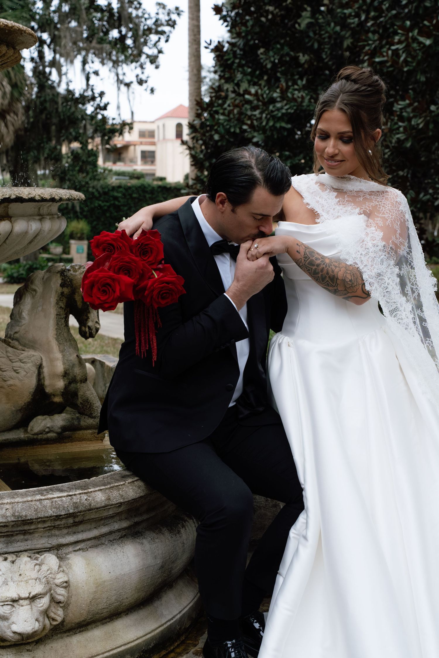 A romantic wedding couple embraces near an ornate stone balustrade in a garden setting.