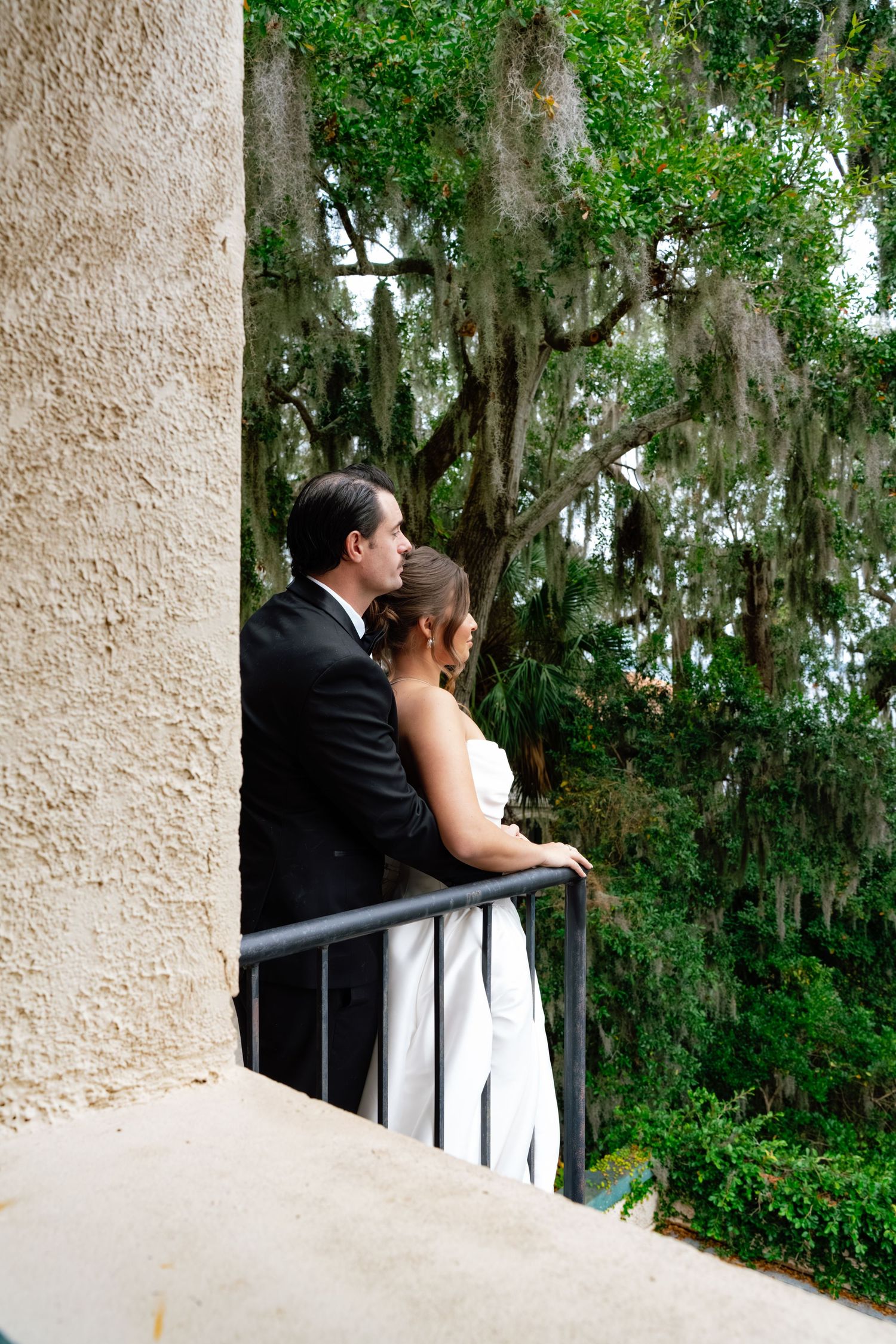 A couple in formal attire share a romantic moment on a balcony overlooking Spanish moss-draped trees.