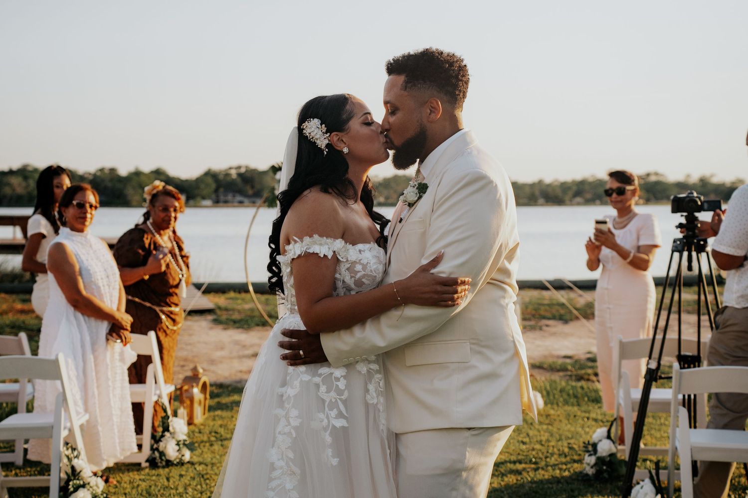A couple shares their first kiss as newlyweds during an outdoor lakeside wedding ceremony at sunset.