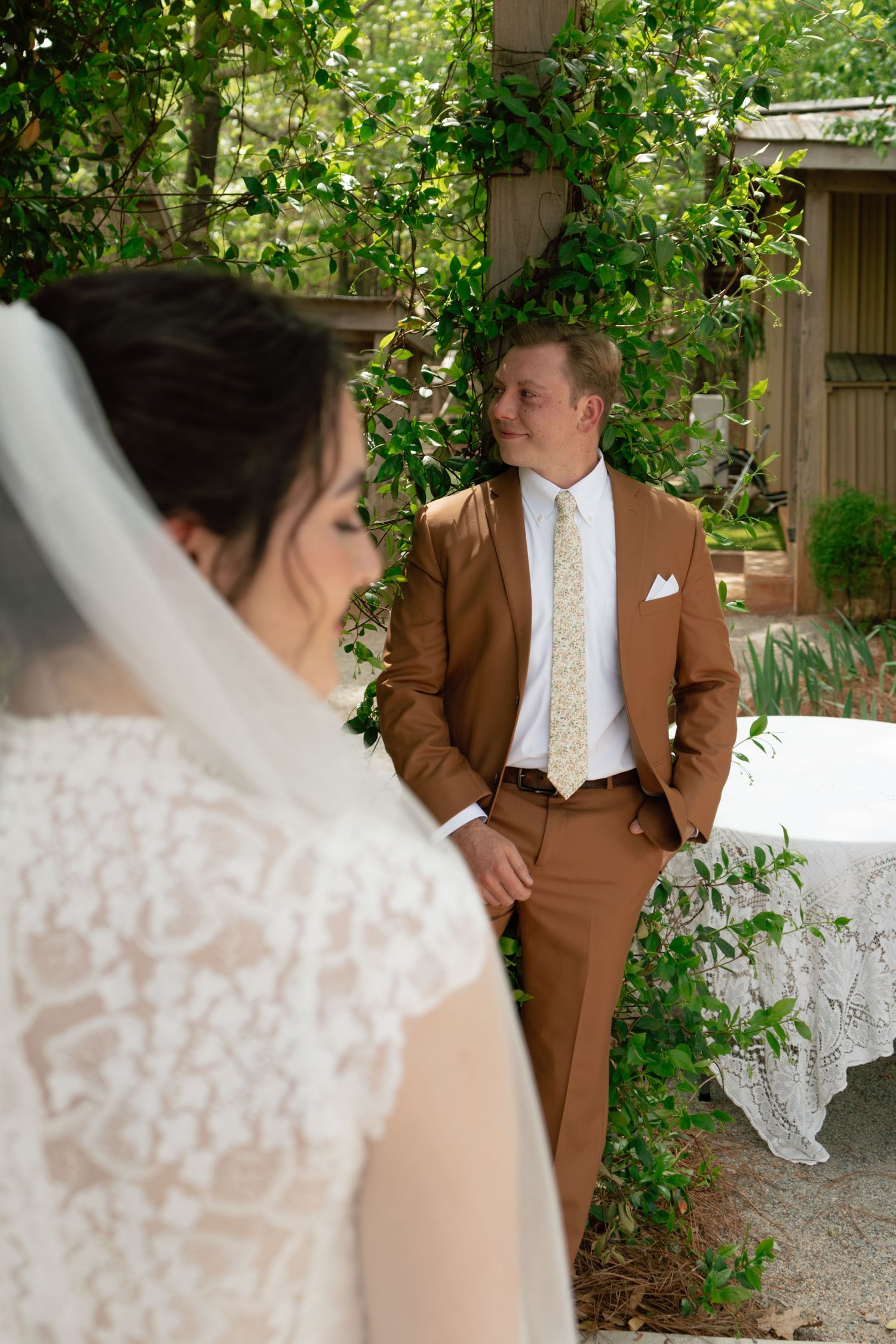 Groom in brown suit smiles at bride in lace wedding dress and veil during outdoor ceremony surrounded by greenery.