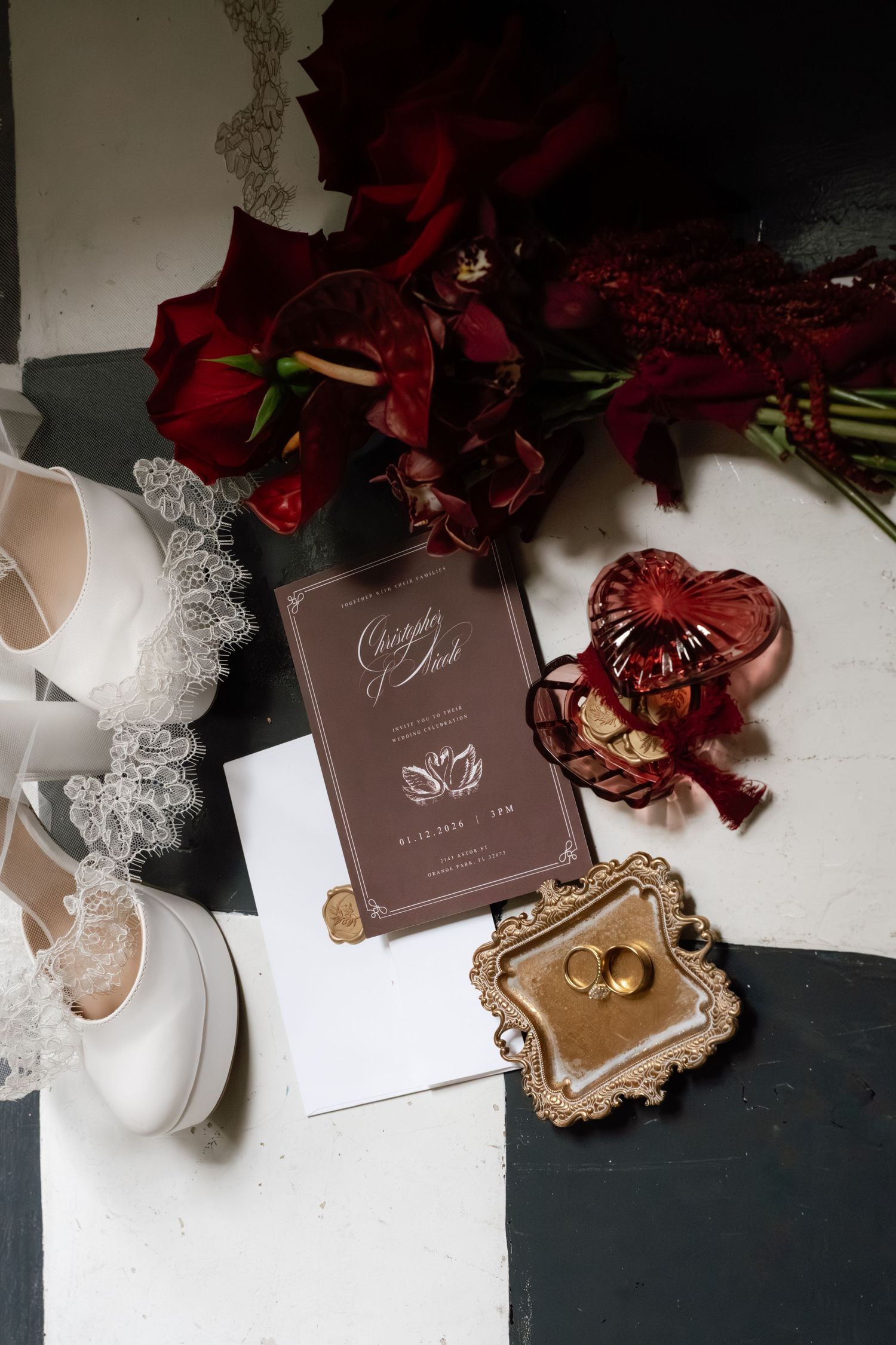 Elegant wedding invitation suite with deep red roses, brown cards, and delicate lace details arranged on a dark surface.