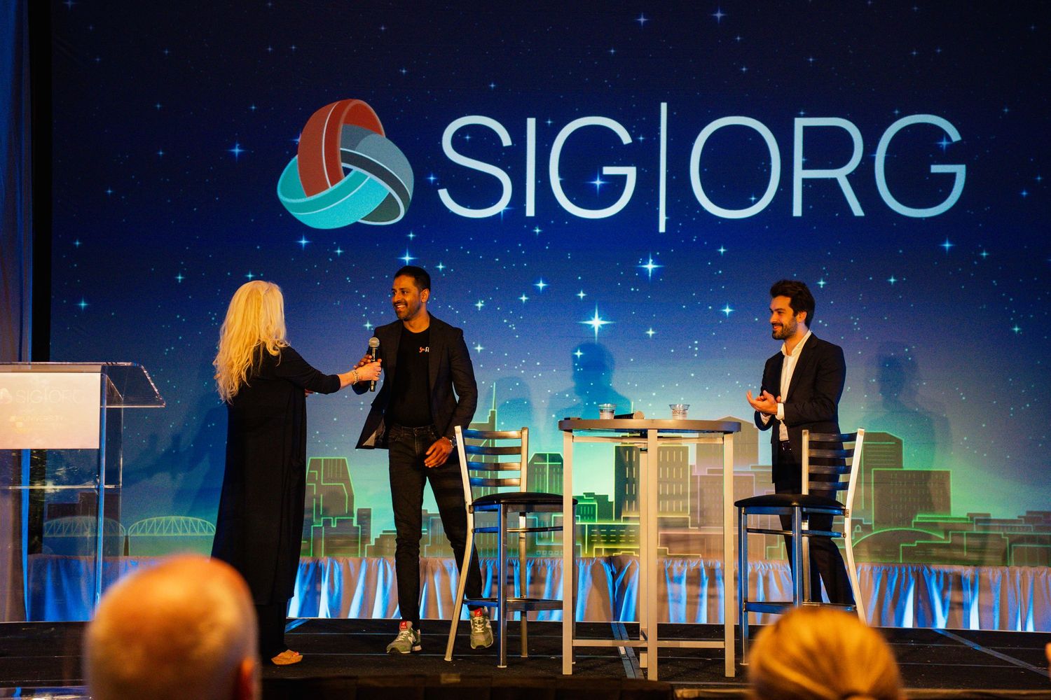 Three professionals networking on stage at a SIG.ORG conference event with a starry blue backdrop.