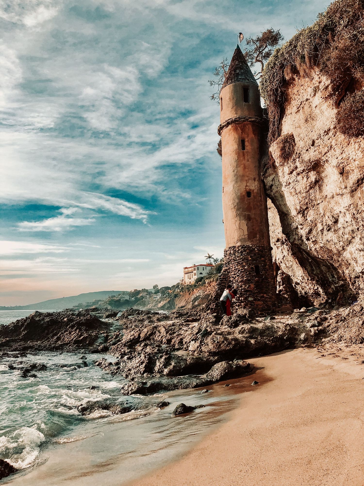 Rustic stone tower perched on a rocky beach cliff with crashing waves and dramatic coastal scenery.