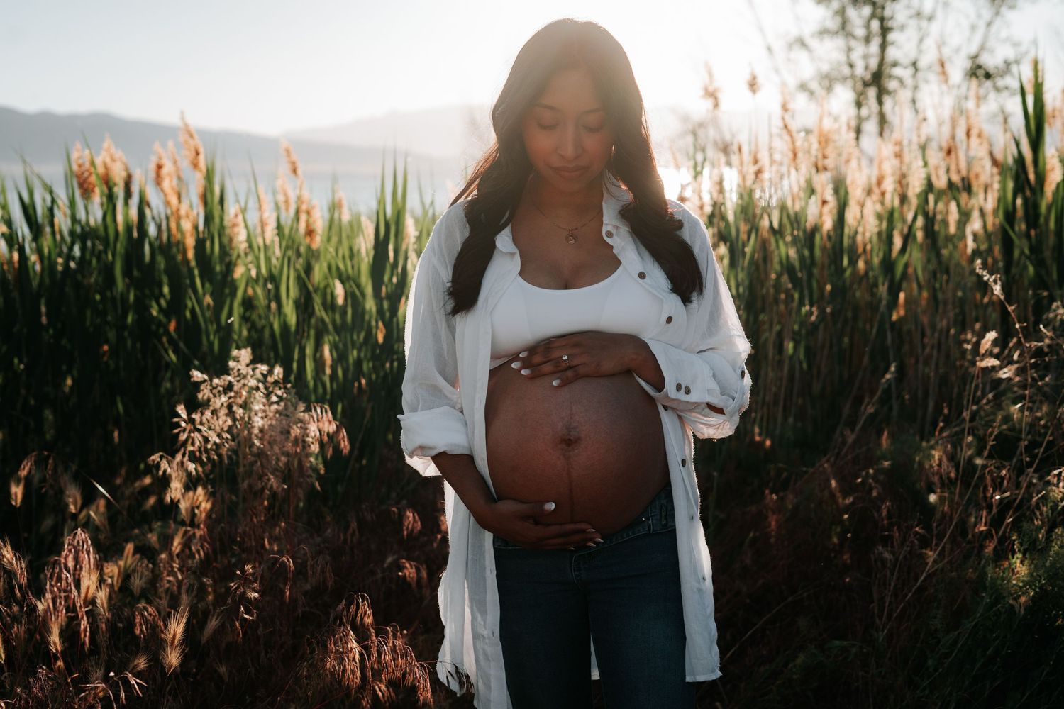 A pregnant woman stands in a field of tall grass and reeds during golden hour sunset.