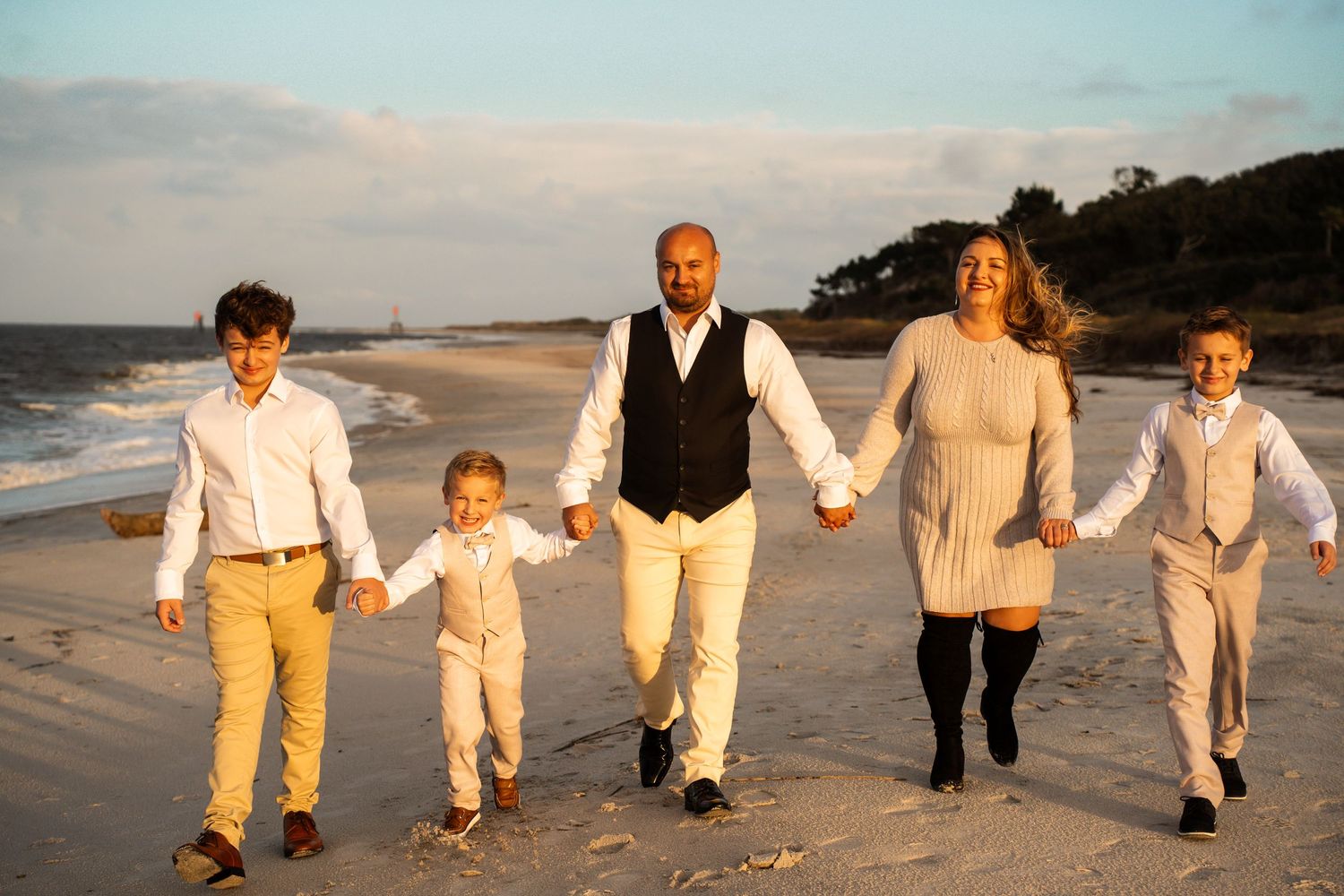 Family strolls along sandy beach at golden hour in matching beige and white outfits.