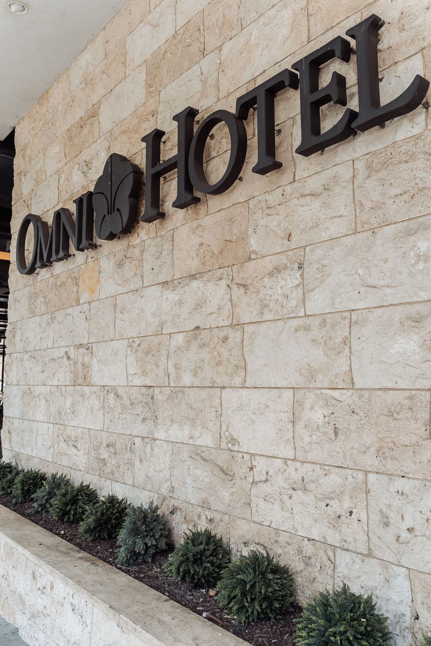 Omni Hotel exterior sign mounted on a elegant beige stone wall with decorative shrubs below.