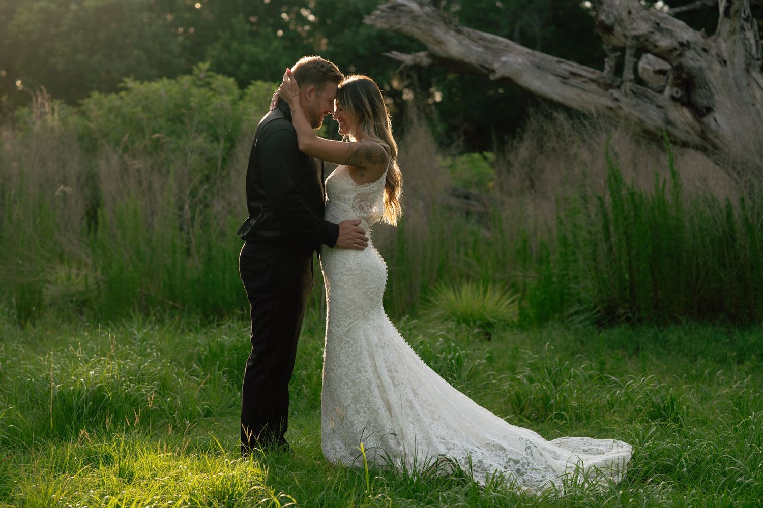 Bride and groom embracing in lush green meadow at golden hour, bride wearing elegant lace mermaid wedding gown.