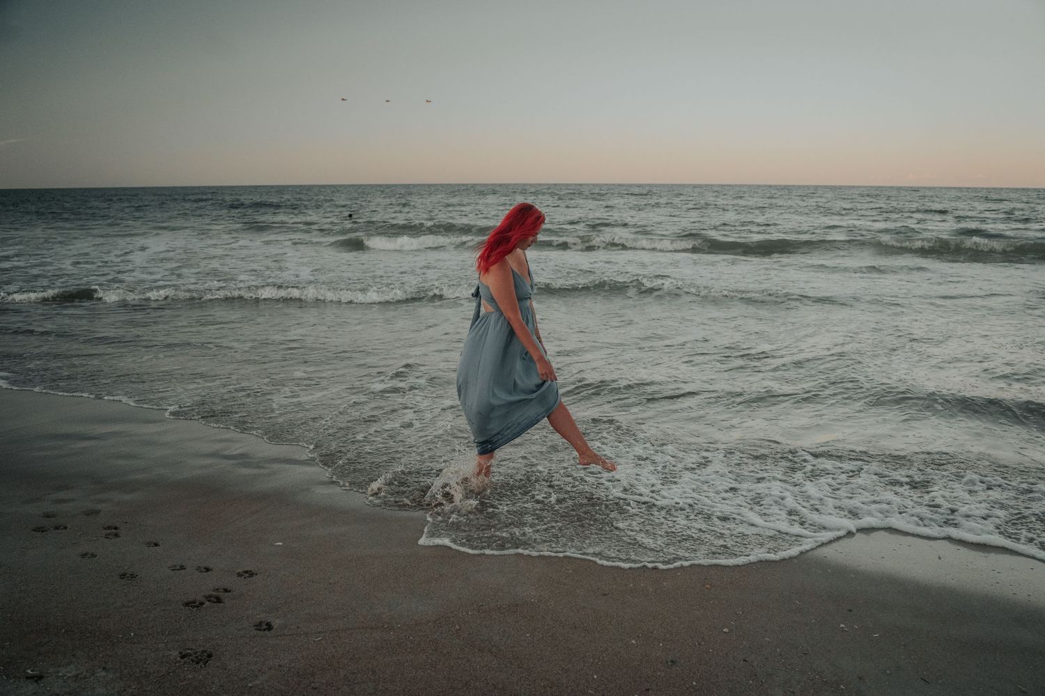 A person with bright red hair in a blue dress splashes through ocean waves at sunset on a sandy beach at Turtle Dunes on Amelia Island