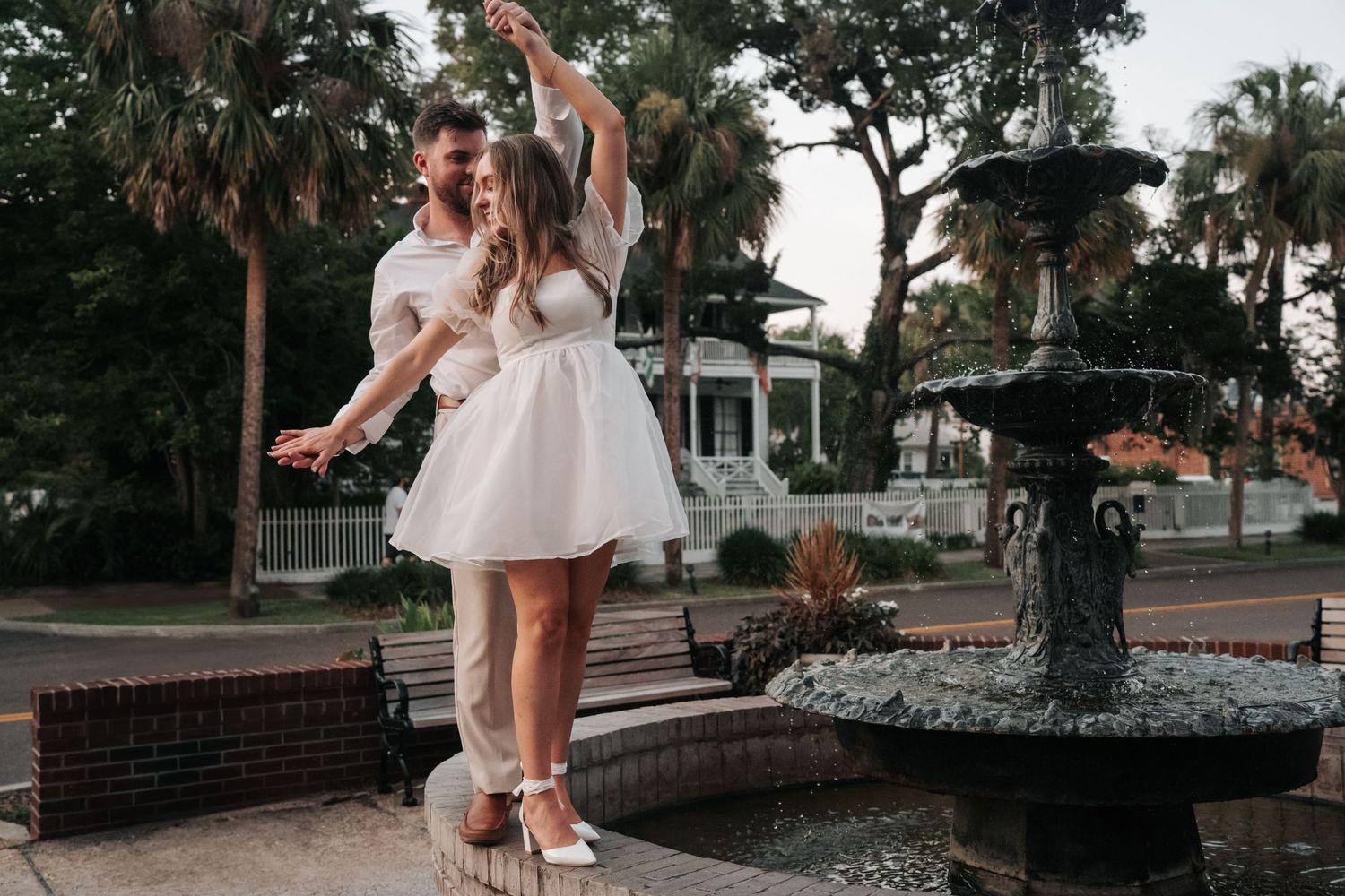 A couple dances together near a fountain in a garden setting at dusk, with palm trees and a white gazebo in the background.